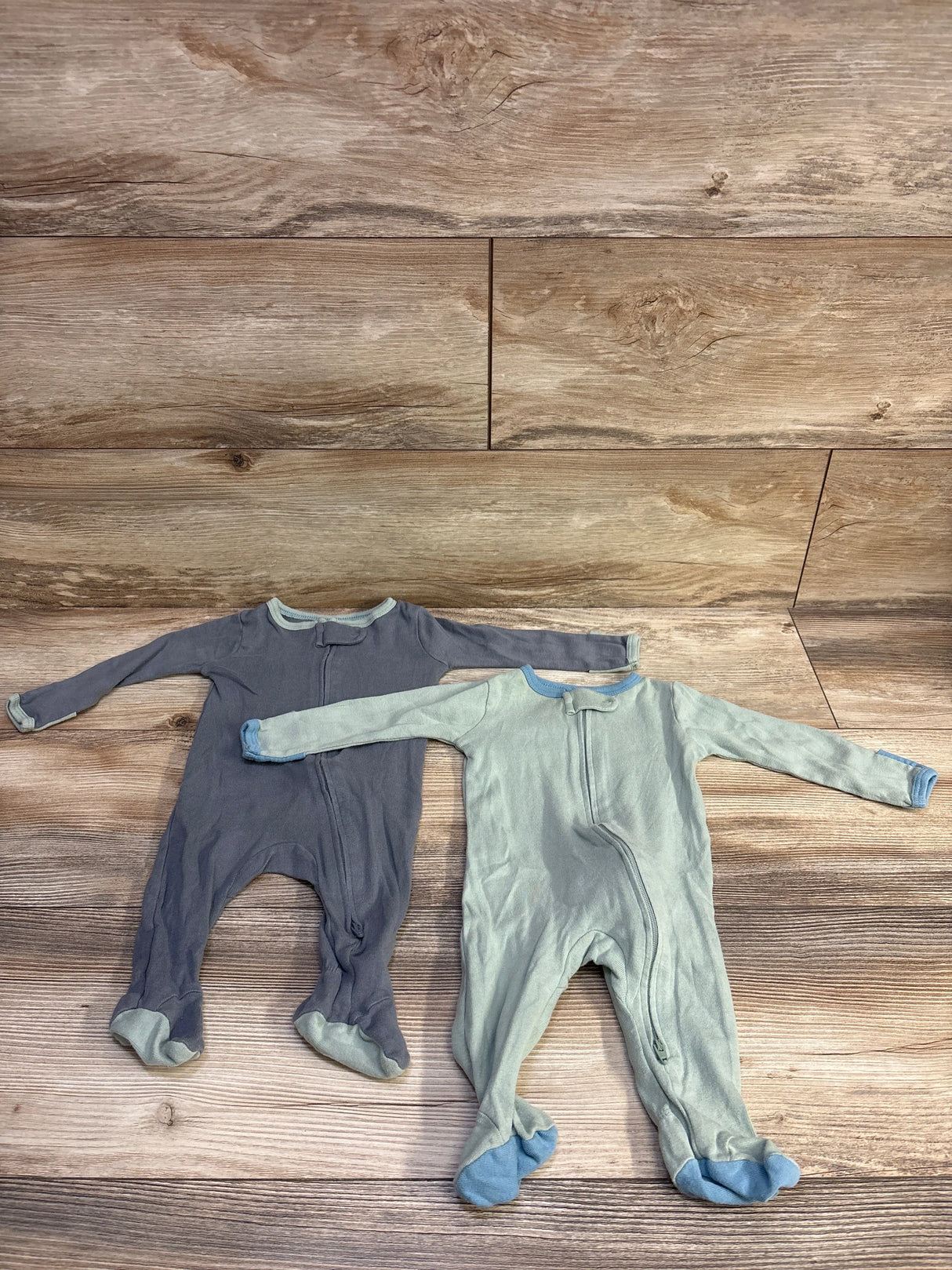 Front view of two Cloud Island footed baby sleepers, green and gray, laid flat showing front zippers on wood background