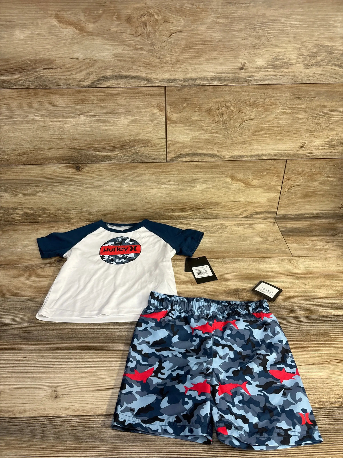 Front view of Hurley toddler 2-piece rashguard set: white shirt with navy sleeves and camo Hurley logo, blue camo shorts with red sharks