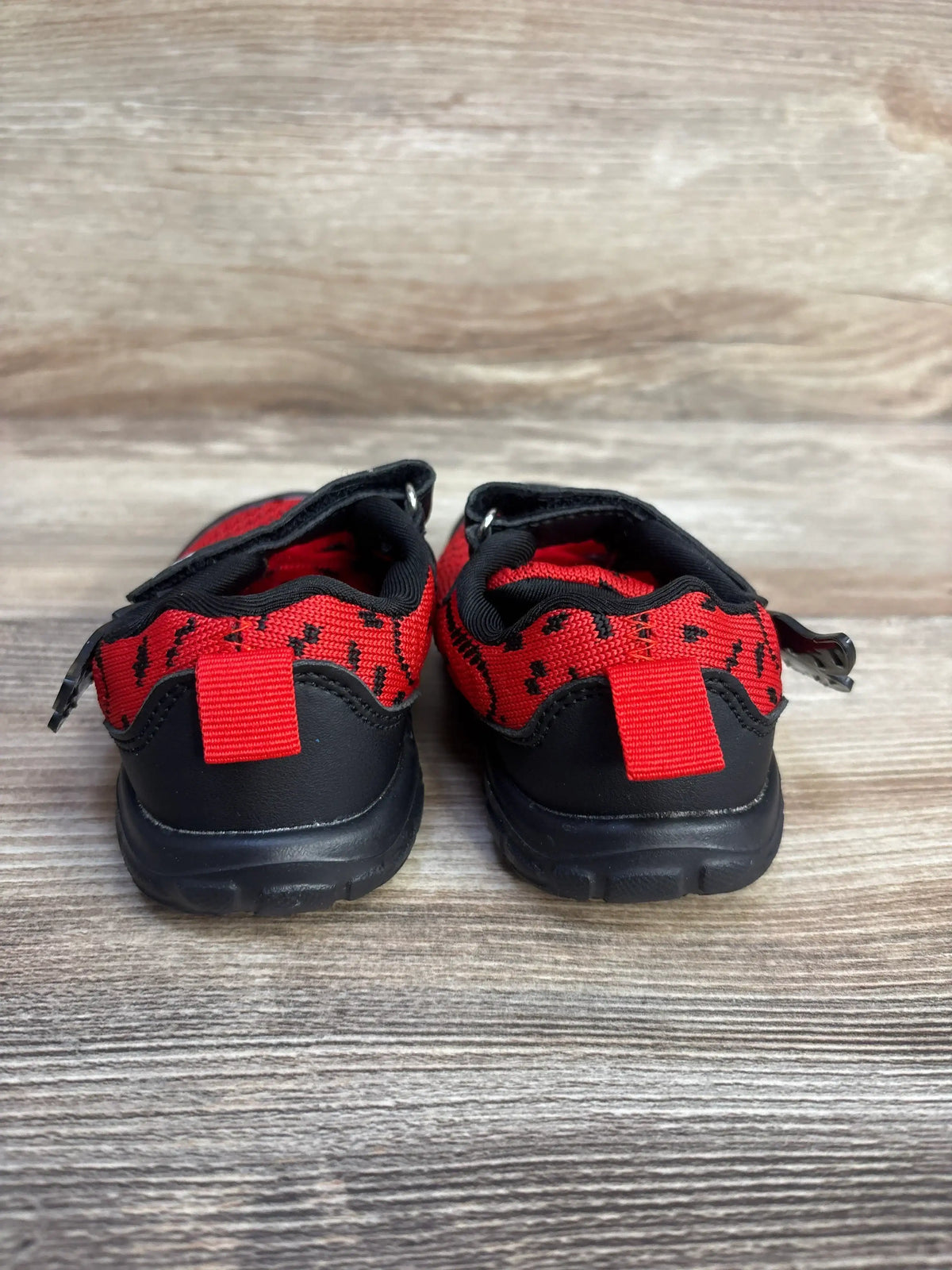 Rear view of boy's red and black wing sneakers showcasing heel pull tabs and black rubber soles on wood plank background