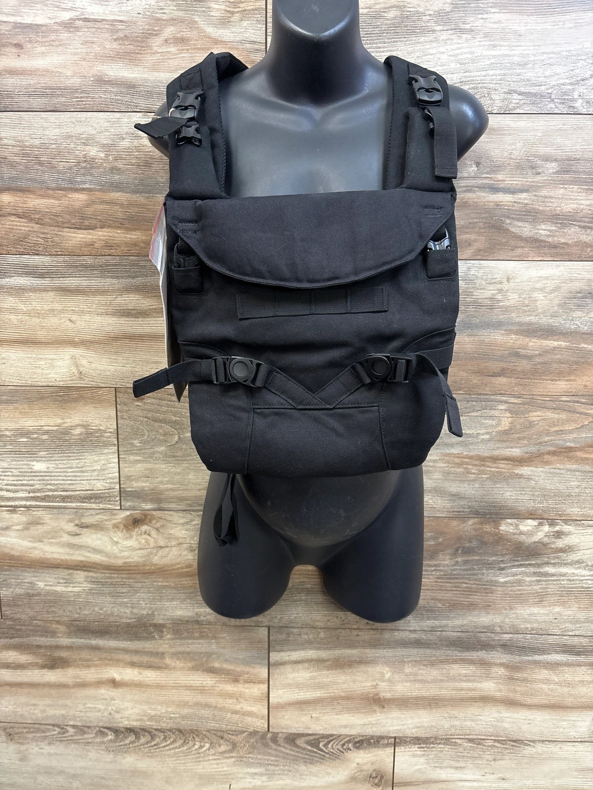 Front view of Tactical Baby Gear carrier in black showcasing padded shoulder straps, front flap and buckles on wood background