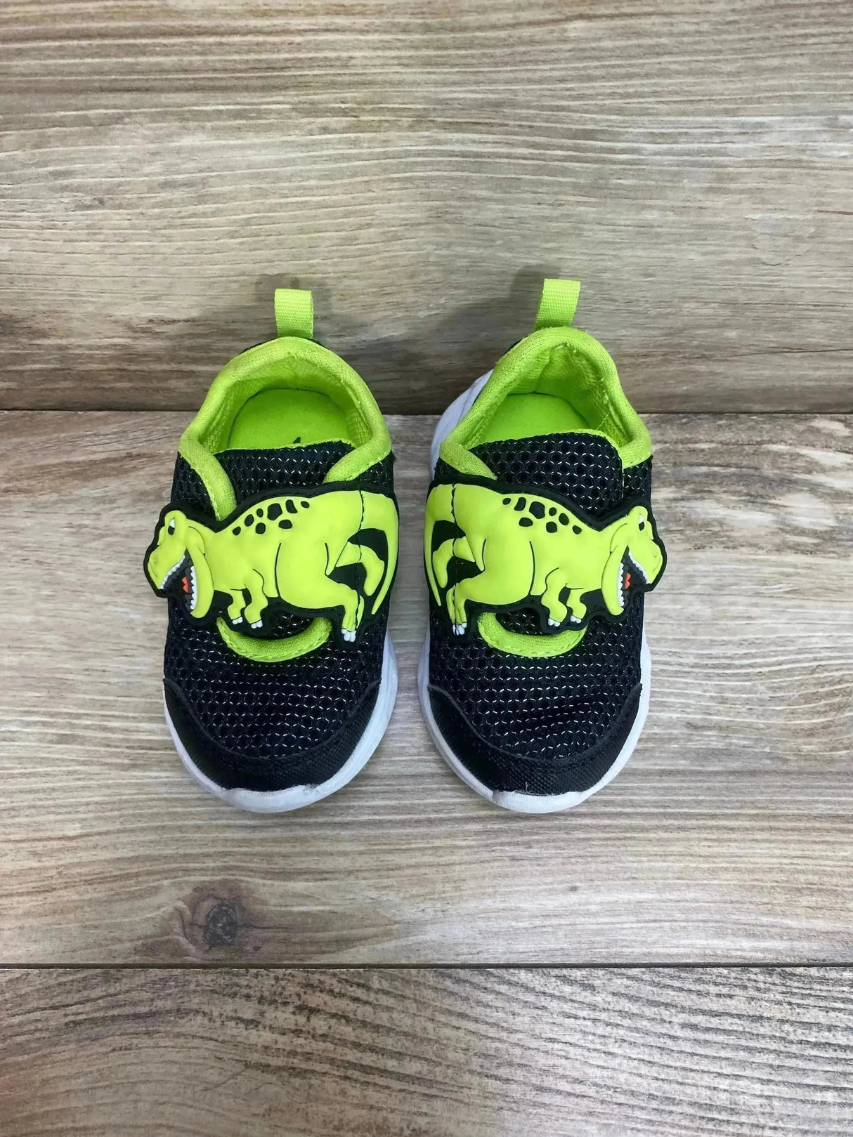 Top-down view of Carter's toddler light-up sneakers showcasing green dinosaur appliques and black mesh upper on wood floor