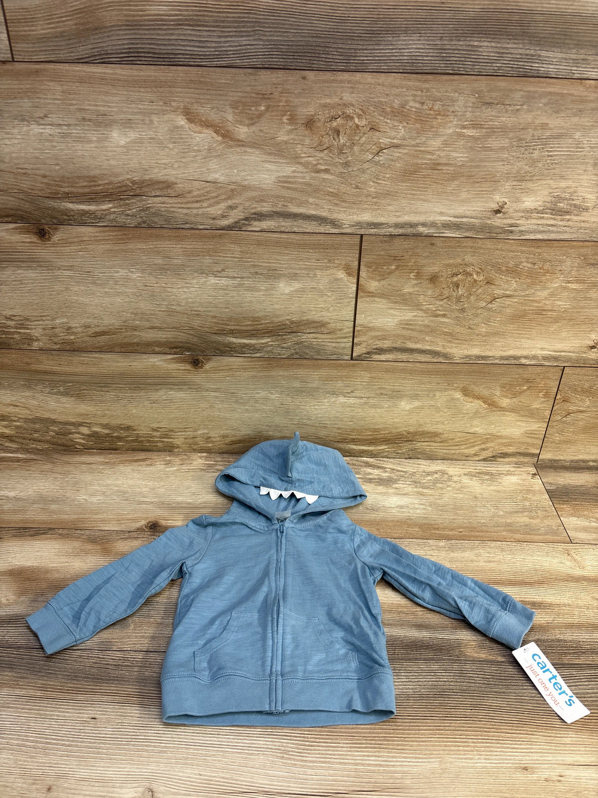 Front flat lay of blue baby shark hooded full zip jacket with white felt teeth on hood and front pockets on wood background
