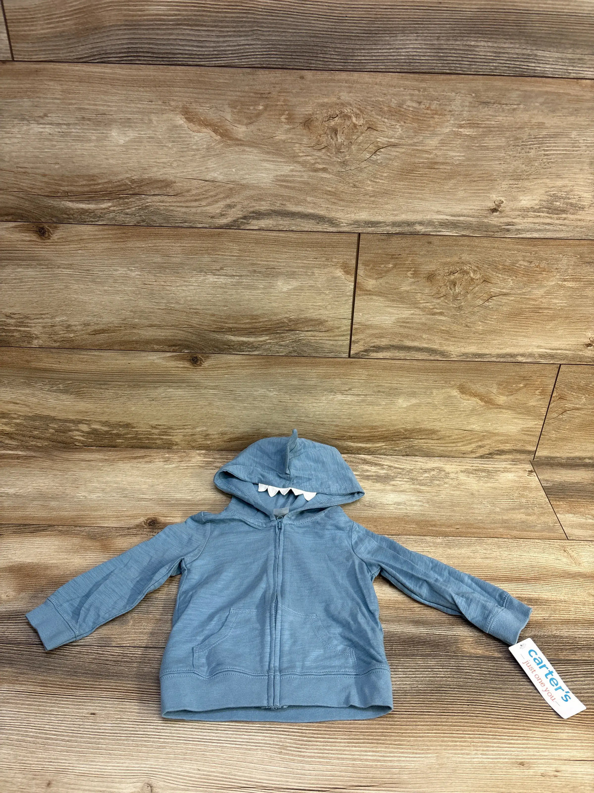 Front flat lay of blue baby shark hooded full zip jacket with white felt teeth on hood and front pockets on wood background