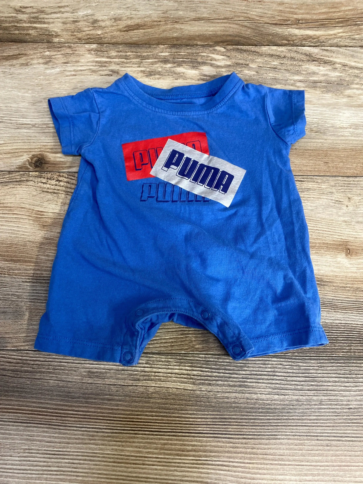 Front view of blue newborn Puma shortie romper showing stacked Puma logo graphics and snap closures at crotch on wood background