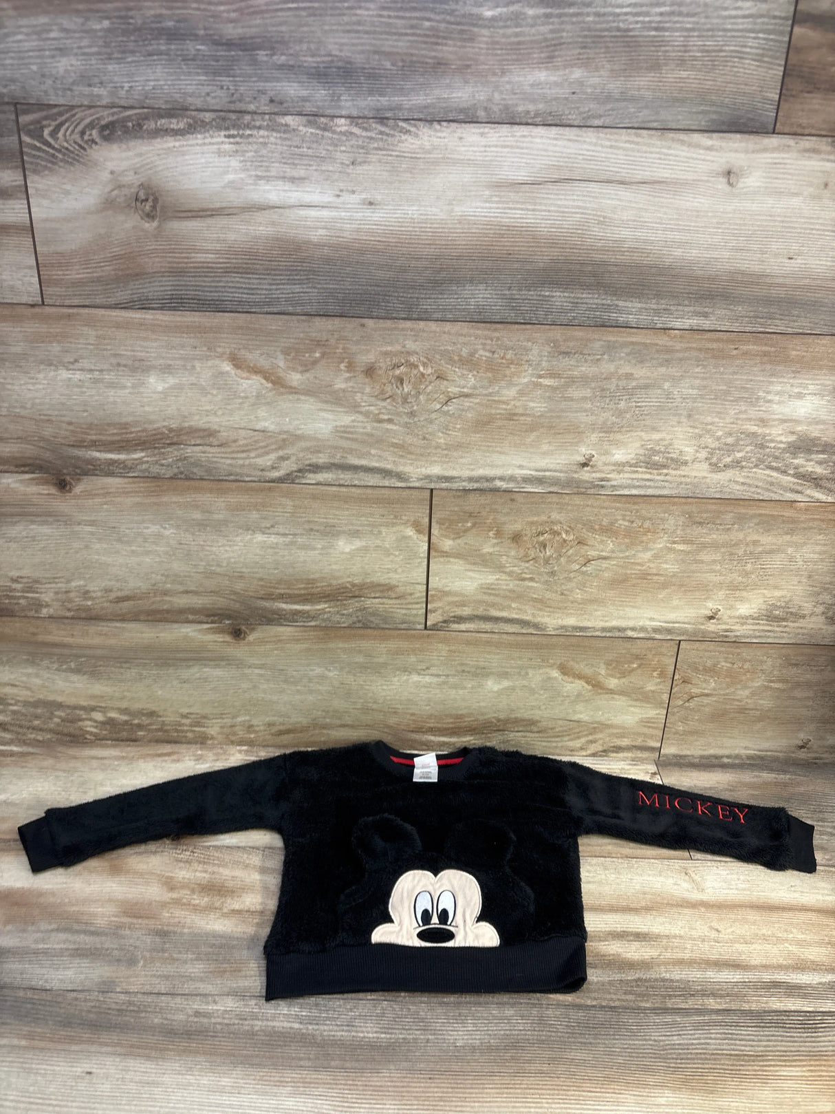 Flat lay of black toddler plush Mickey Mouse pullover sweatshirt showing front Mickey face applique and red MICKEY sleeve text on wood floor