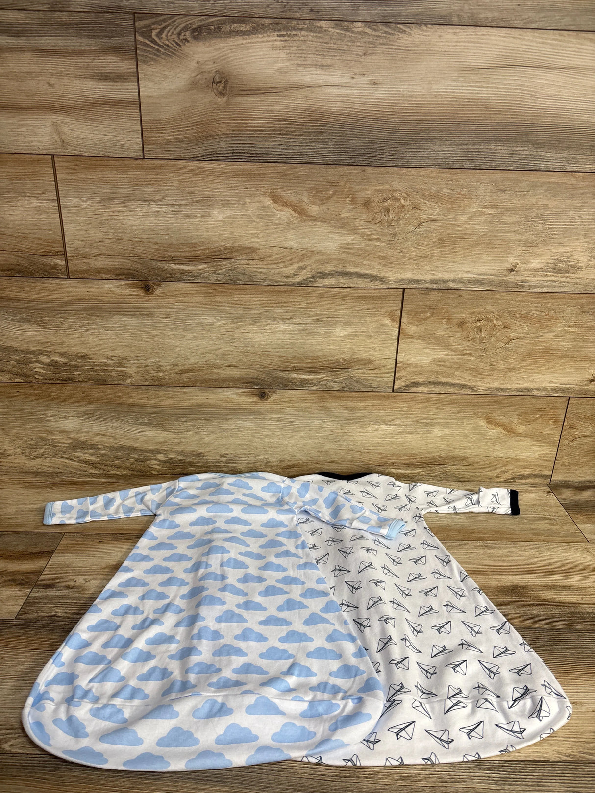 Two baby sleep sacks with long sleeves laid on wooden floor, one blue clouds, one black paper planes pattern