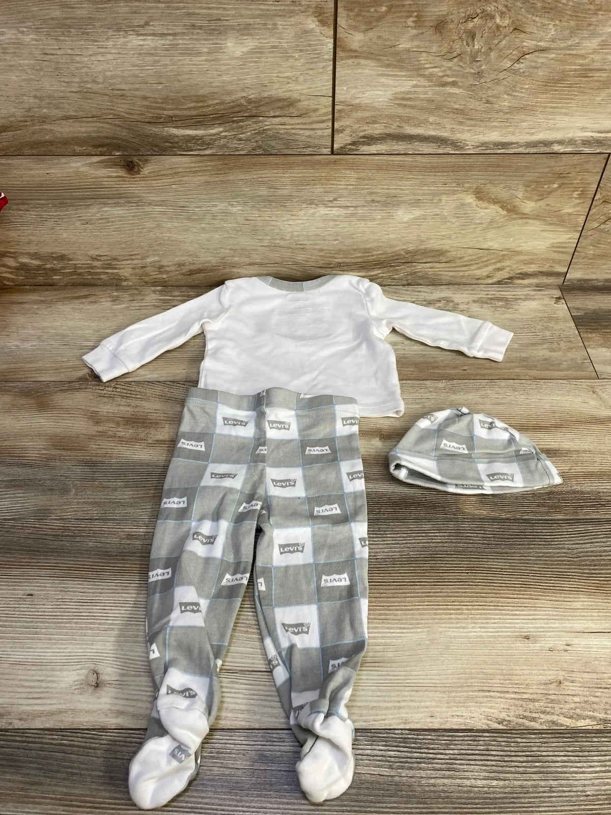 Flat lay of Levi’s 3-piece baby outfit 6M: white long-sleeve top, gray footed pants with Levi’s logo, matching hat on wood