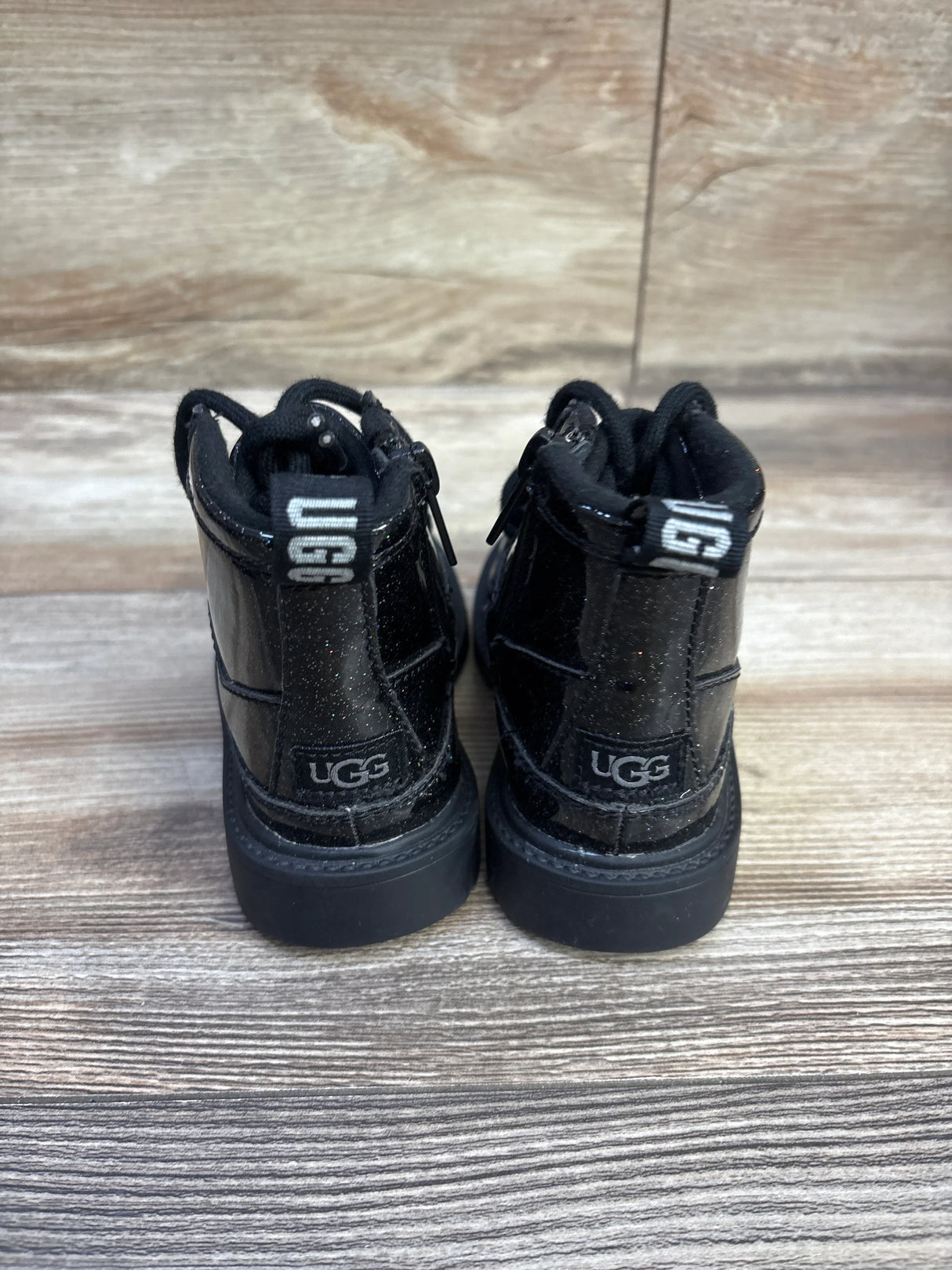 Rear view of UGG Kids Robley glitter black boots size 7C showcasing glitter finish, UGG heel logo and pull tab on wood background