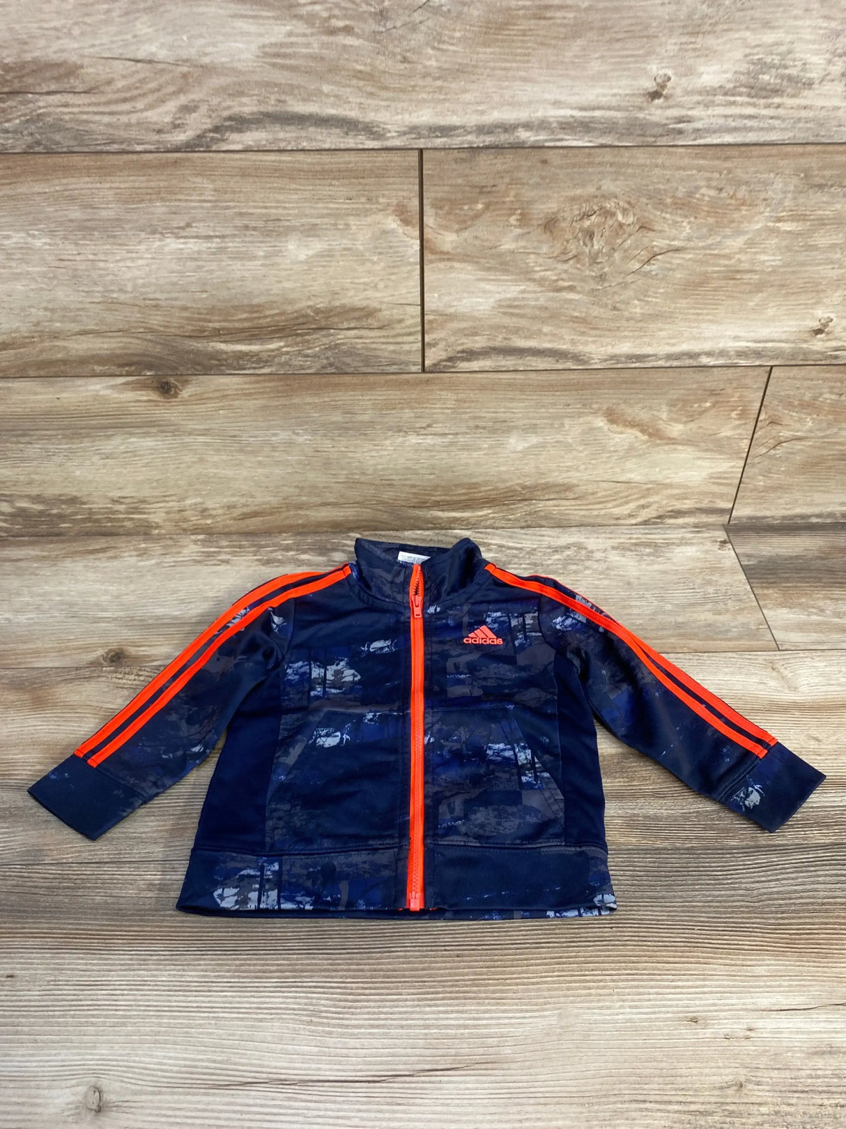 Flat lay front of toddler Adidas navy jacket with bright orange zipper and orange shoulder stripes on wood floor