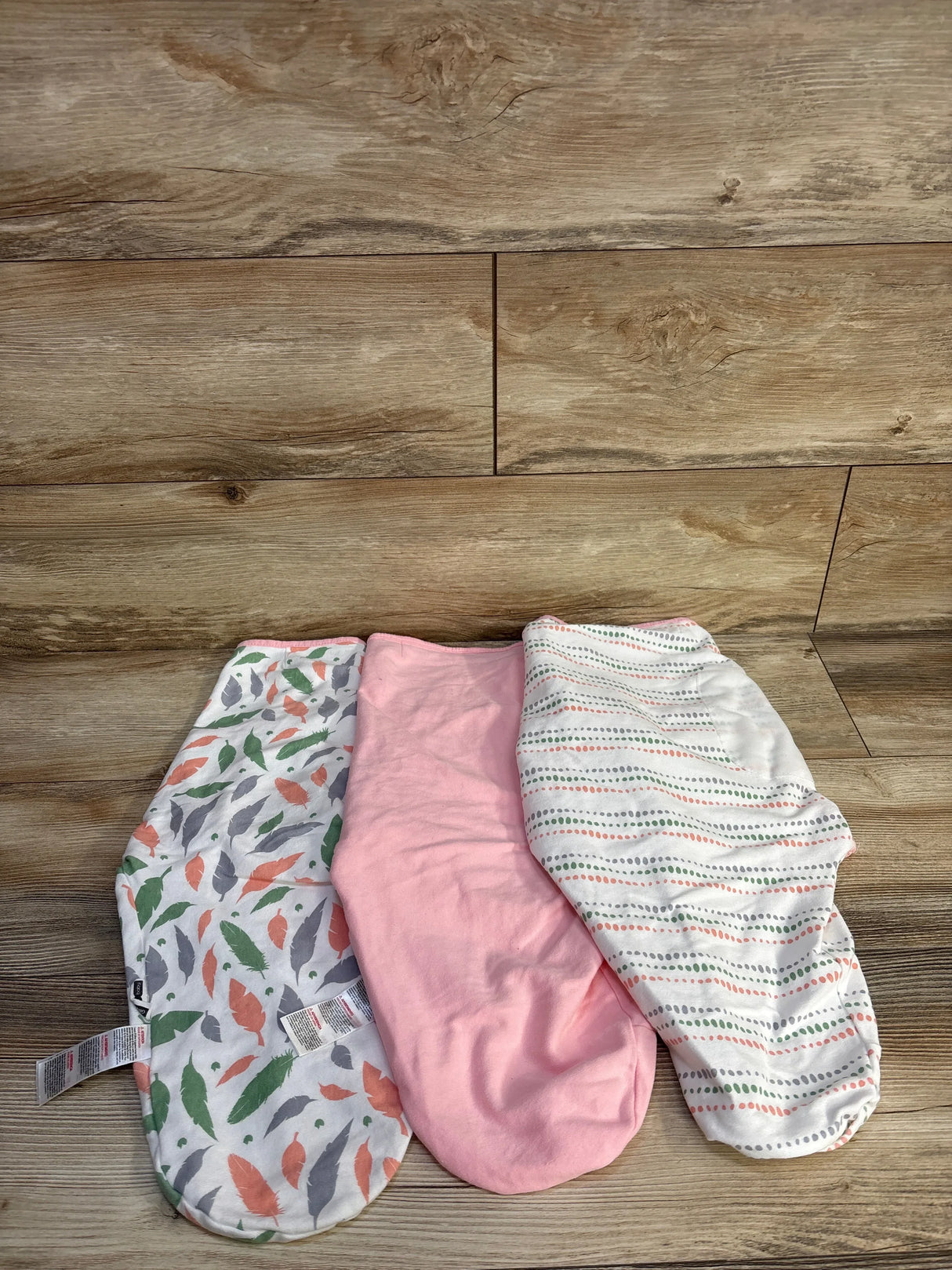 Top-down view of three Gilquen swaddle wraps (feather print, solid pink, dotted stripe) laid flat on wood background, size 3-6 months