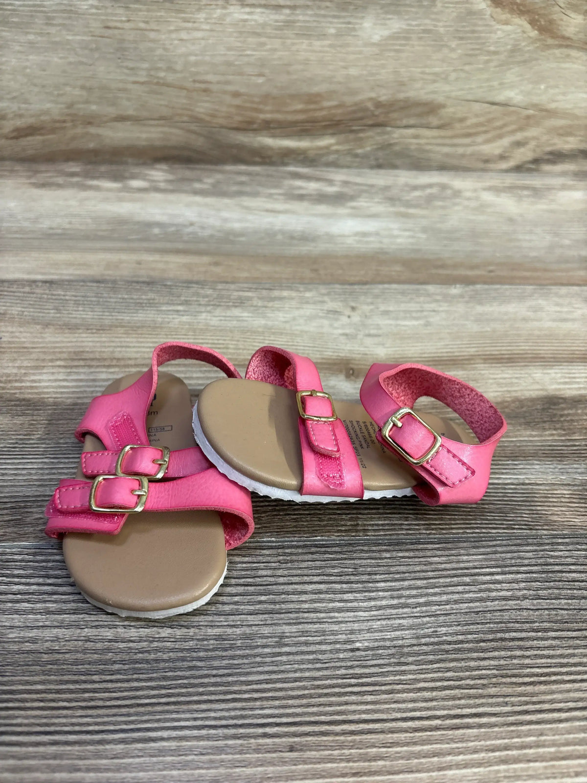 Angled view of Baby Gap pink buckle sandals showcasing two adjustable gold buckles and ankle strap on wood background