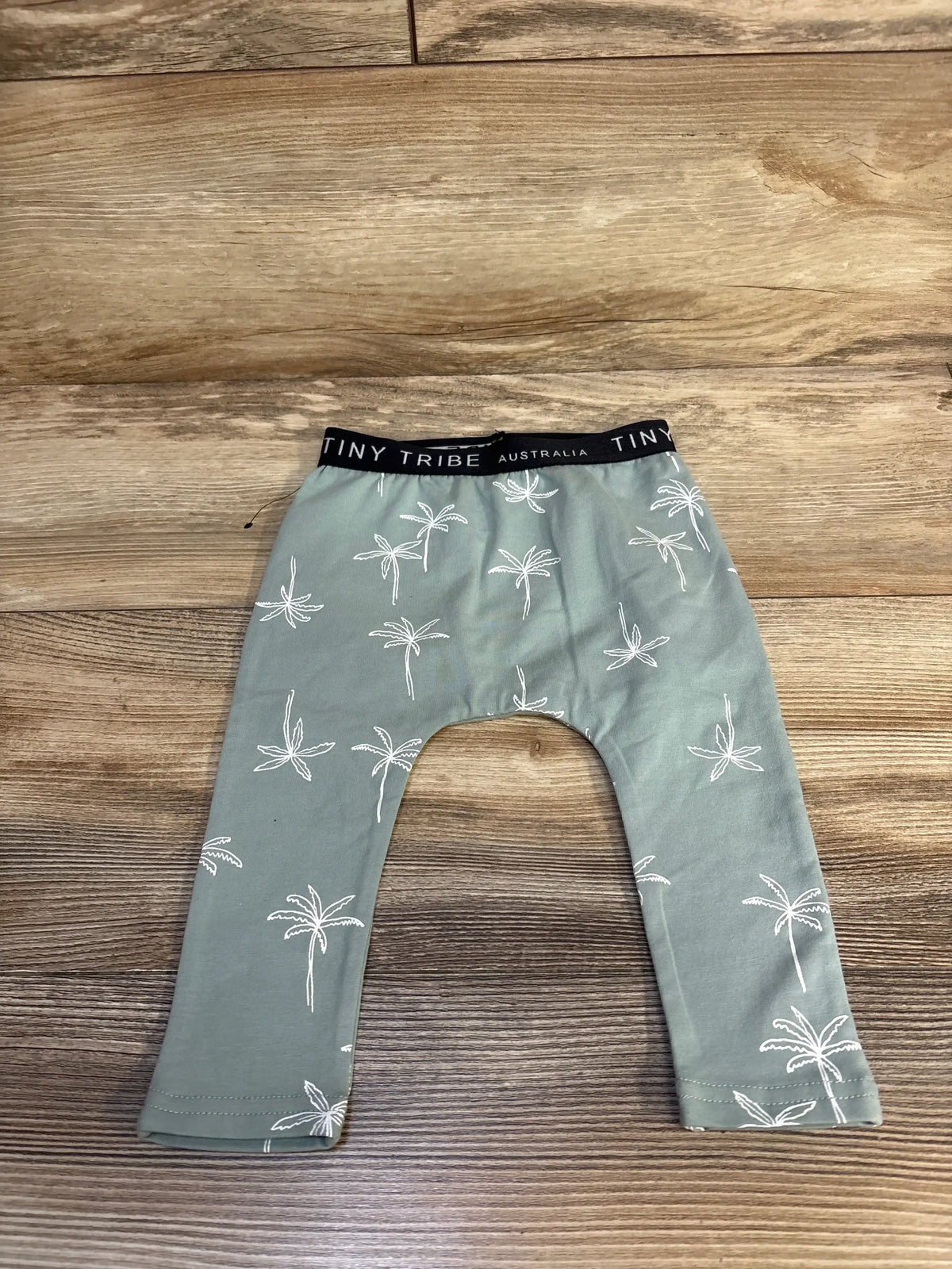 Flat lay of Tiny Tribe pants in sage green with white palm tree print and black waistband labeled TINY TRIBE AUSTRALIA on wood floor