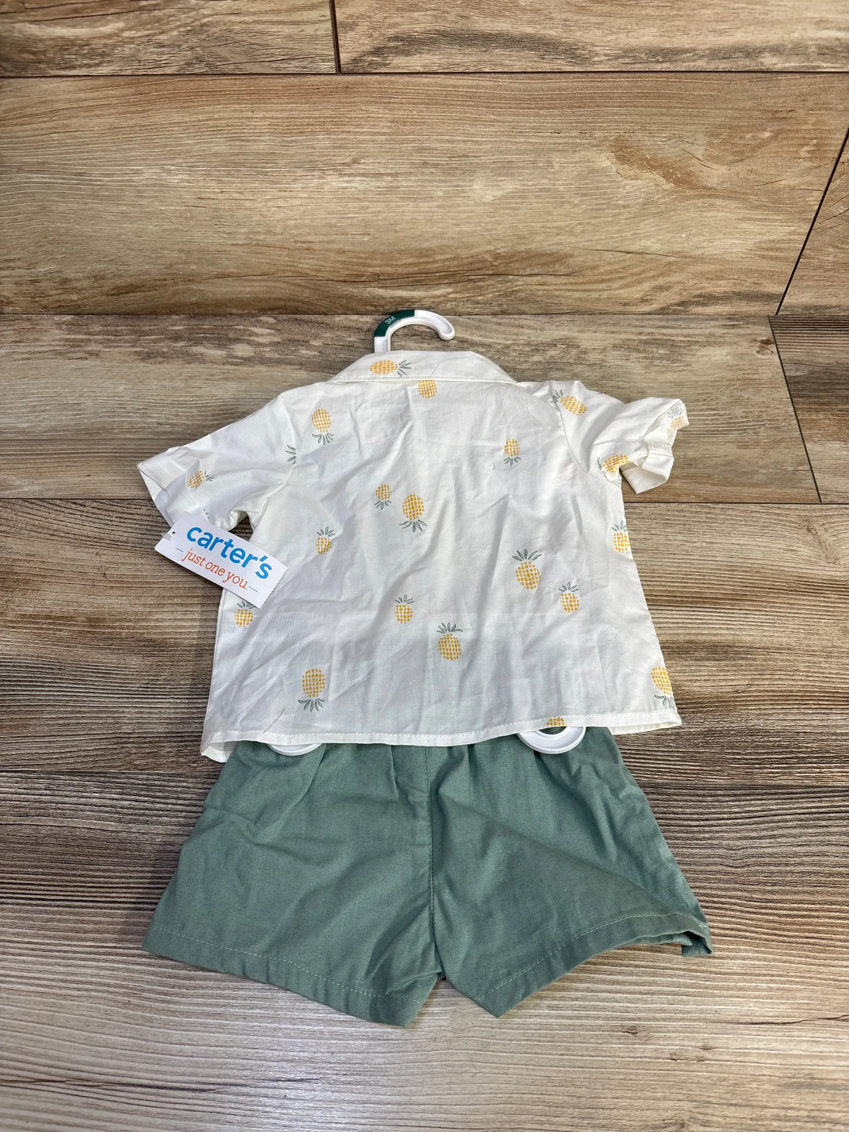 Flat lay of white pineapple-print button-up shirt and green shorts set with Carter’s tag on wood floor