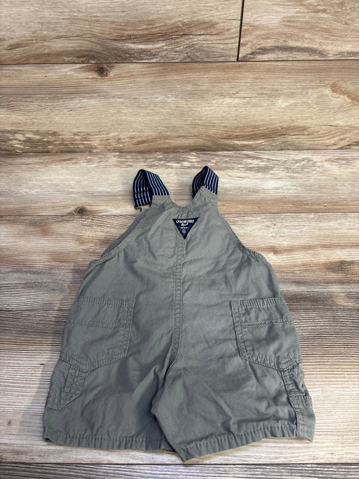 Flat lay of green Oshkosh shortalls, size 18 months, with striped navy straps and front patch pockets on wood background