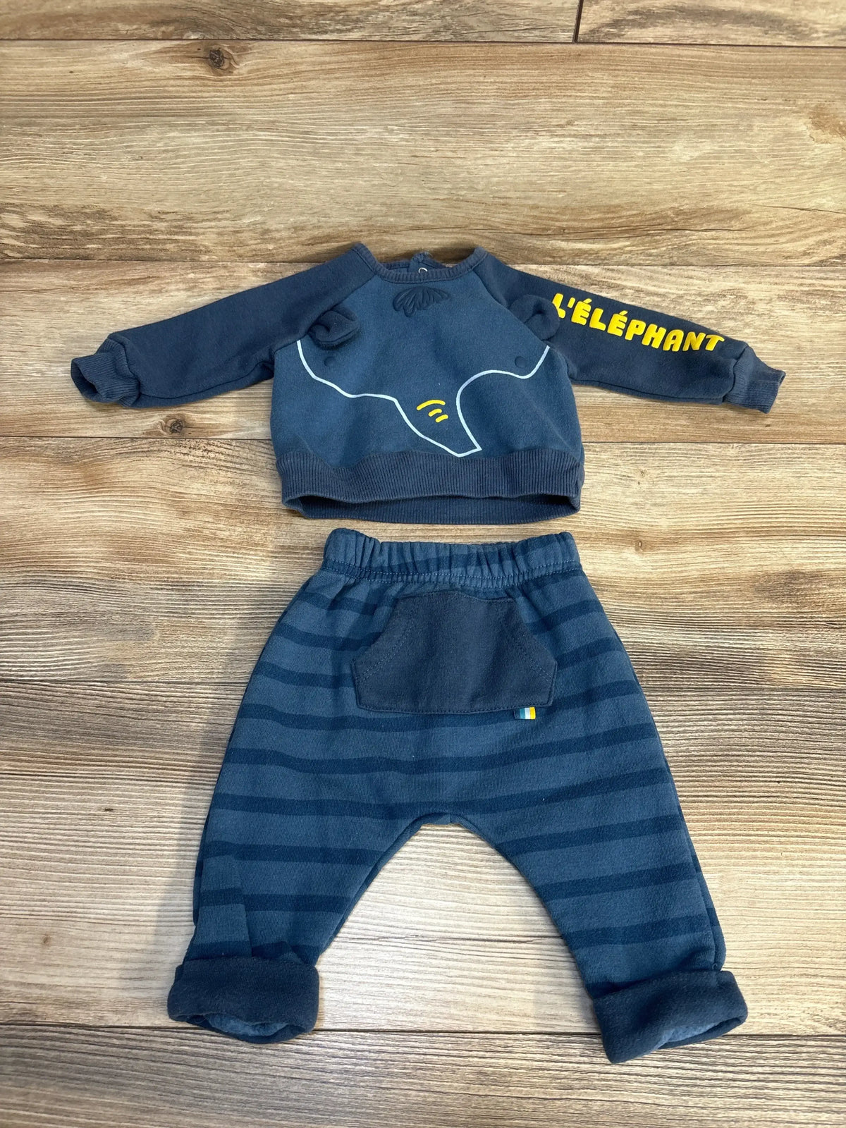 Flat lay of newborn Lily & Jack navy 2-piece sweatshirt set showing elephant outline and yellow sleeve text with striped pants on wood