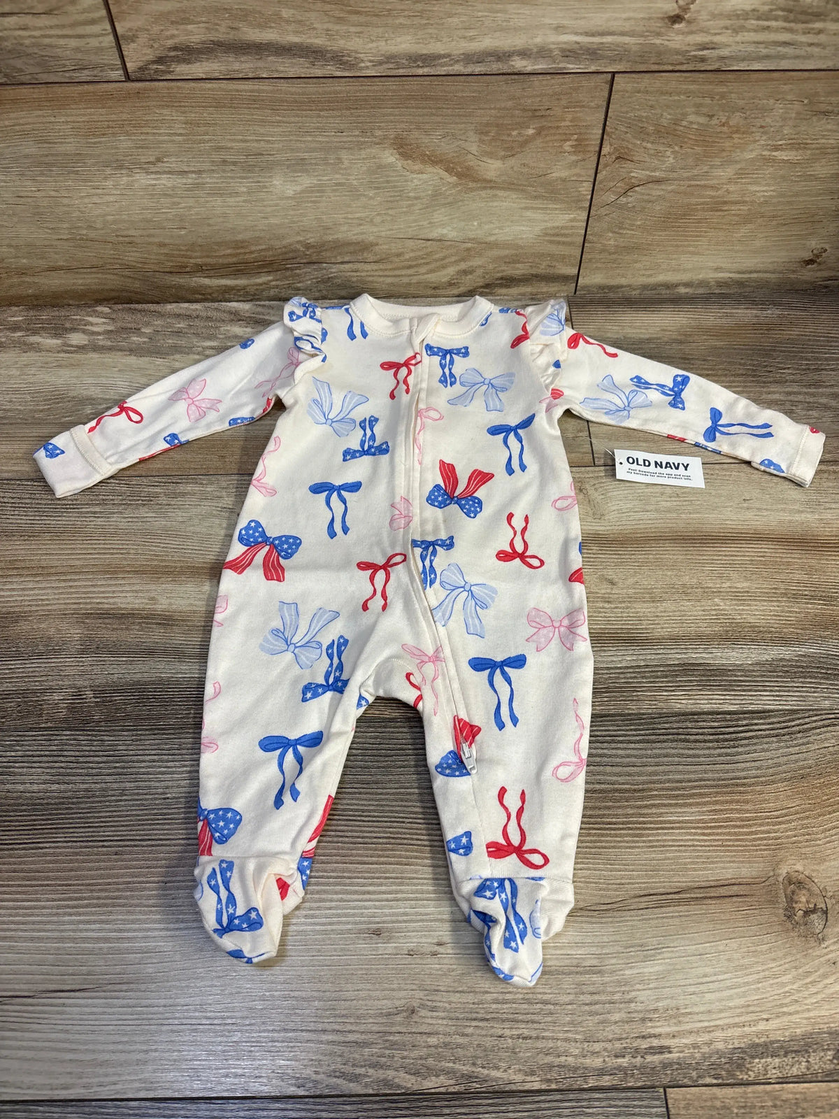 Front view of a cream Old Navy patriotic bow sleeper in size 0 to 3 months, showcasing footed legs on a wood floor