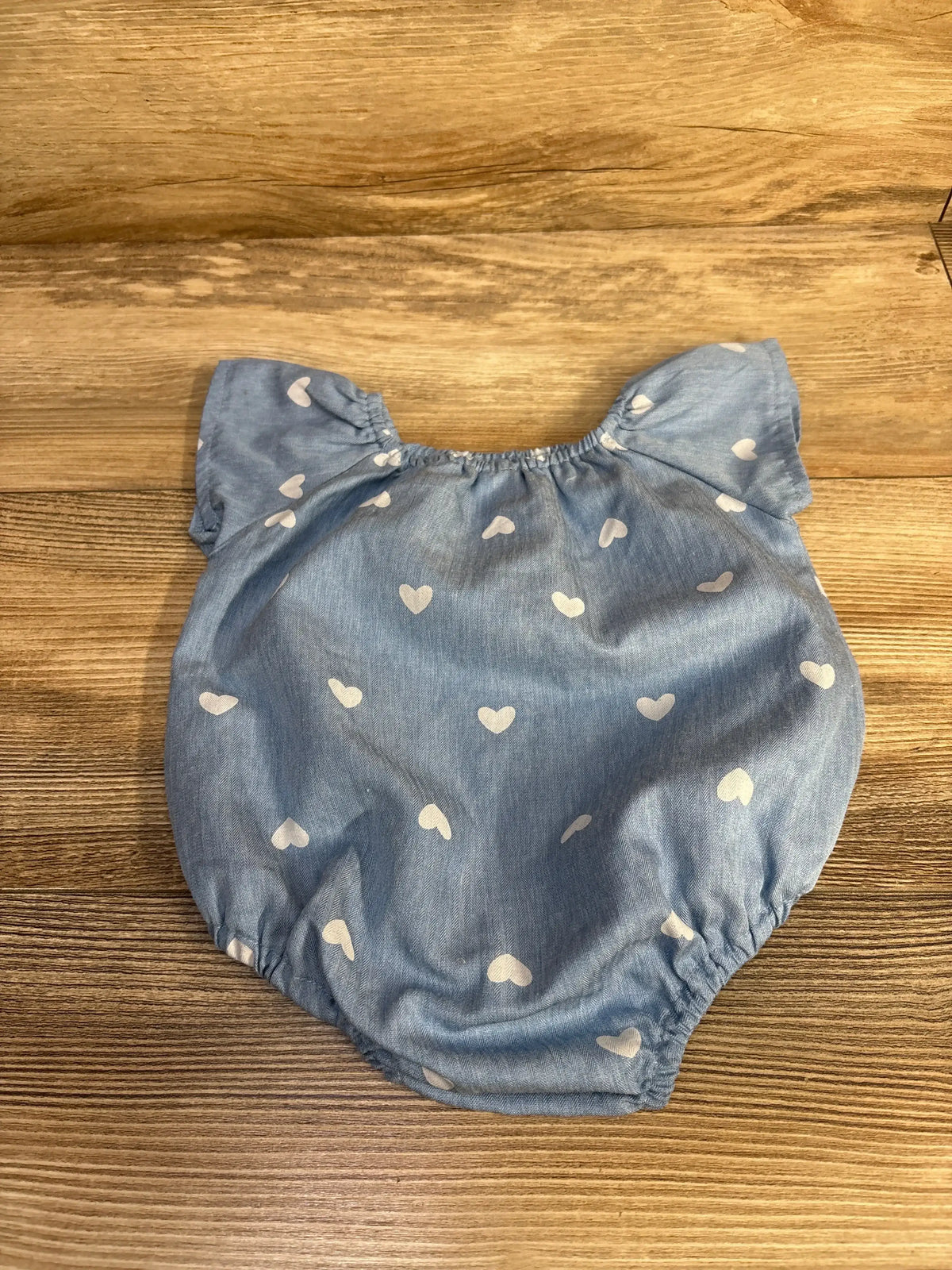 Top-down view of blue baby romper with white heart print (3M), elasticized neckline and leg openings on wood background