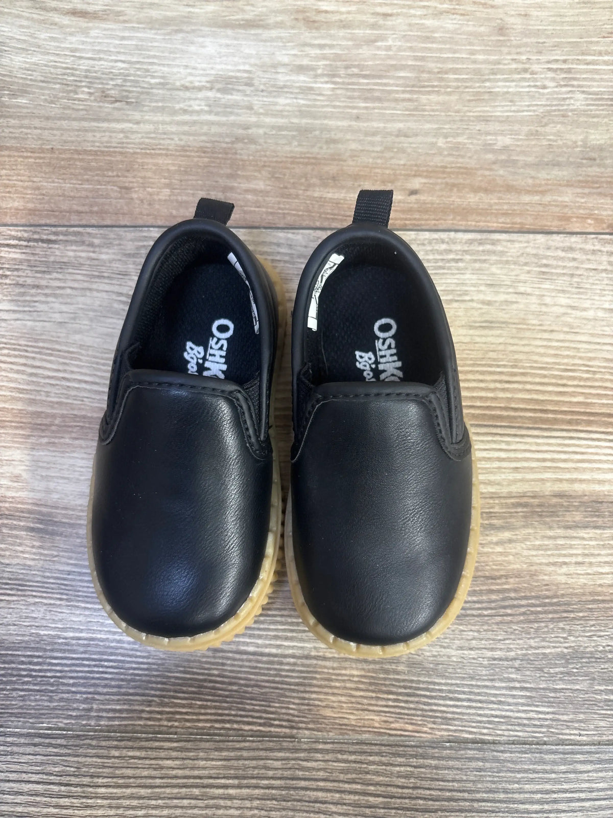 Top view of OshKosh Victor slip-on sneakers in black, size 4C, showcasing pull tabs and translucent soles on a wood-grain background