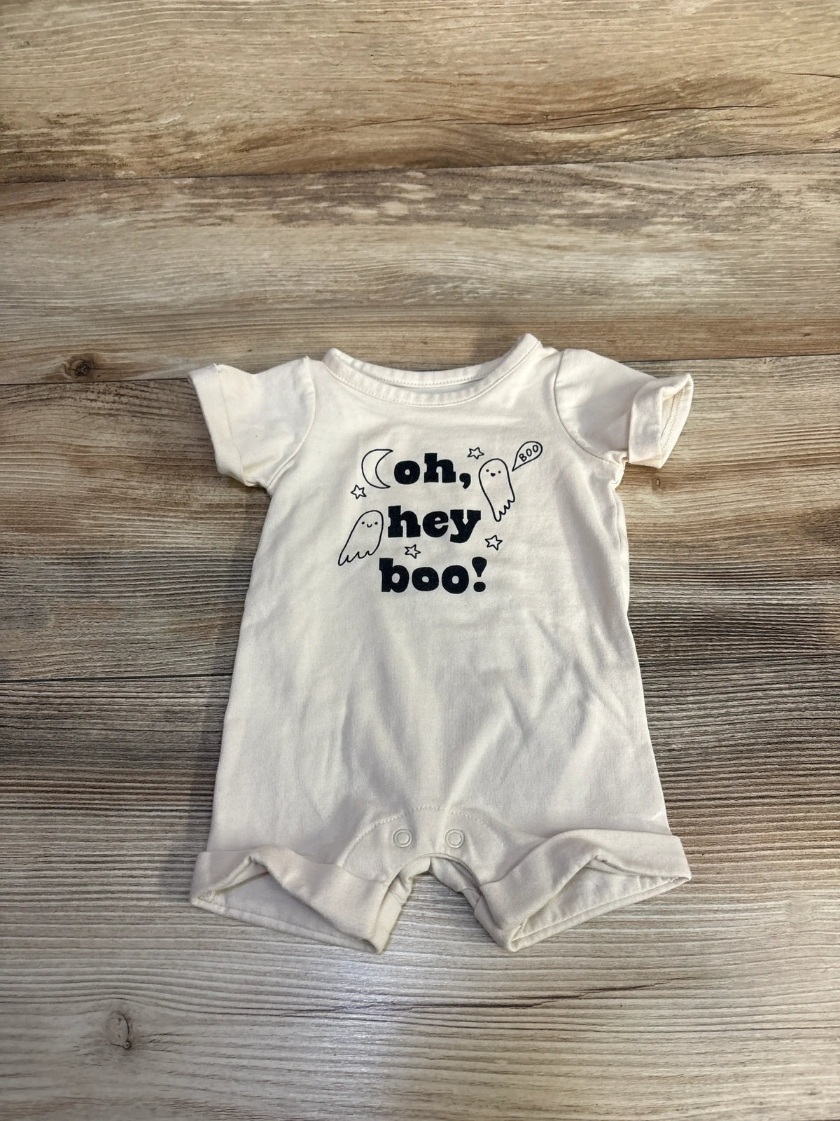 Front view of Cat & Jack newborn shortie romper with ivory fabric, Oh, Hey Boo! ghost print, and snap closures on wood