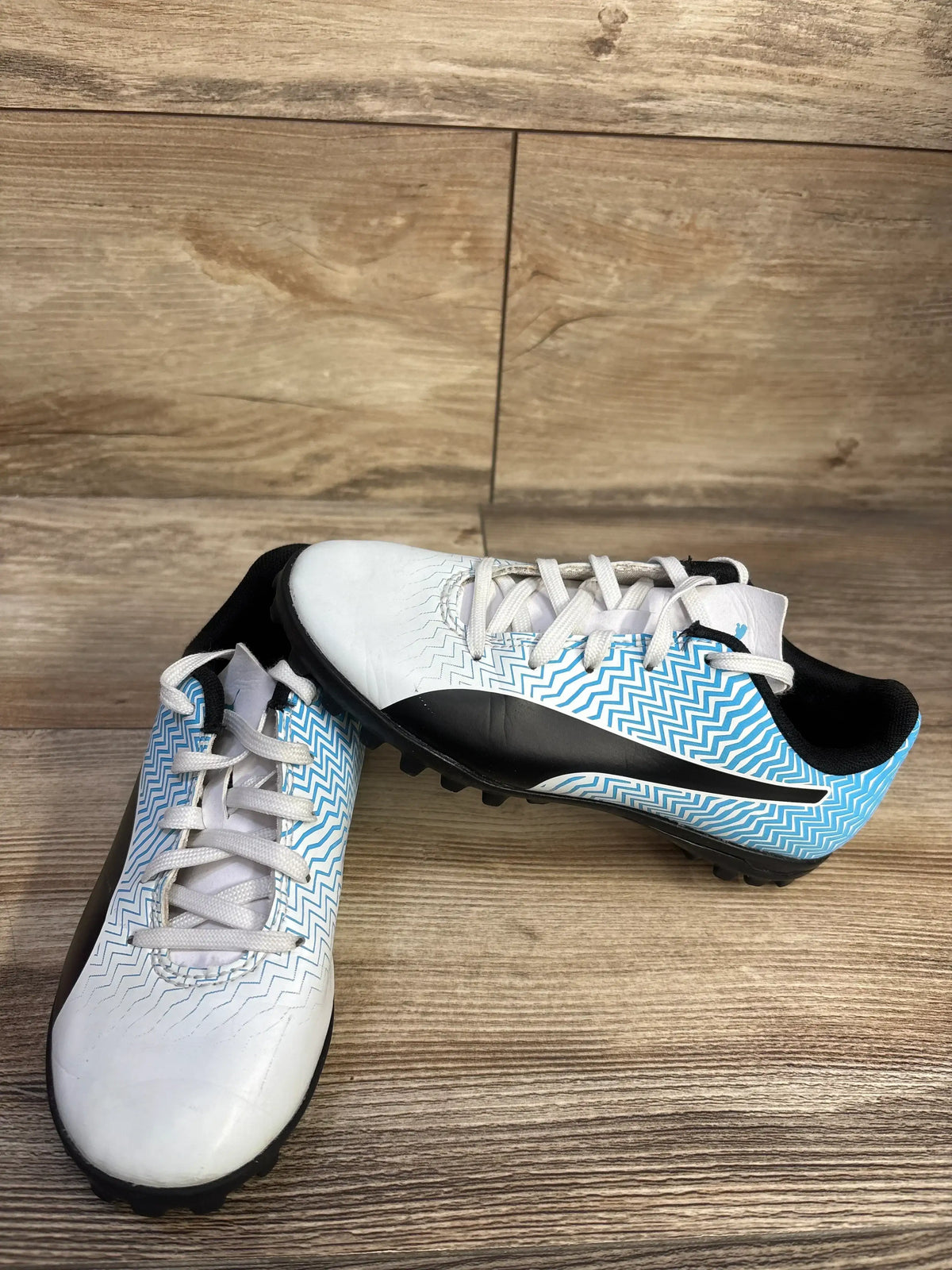 Three-quarter view of kids’ Puma Rapido II soccer cleats in white and blue showing zigzag pattern and black turf-studded sole on wood background