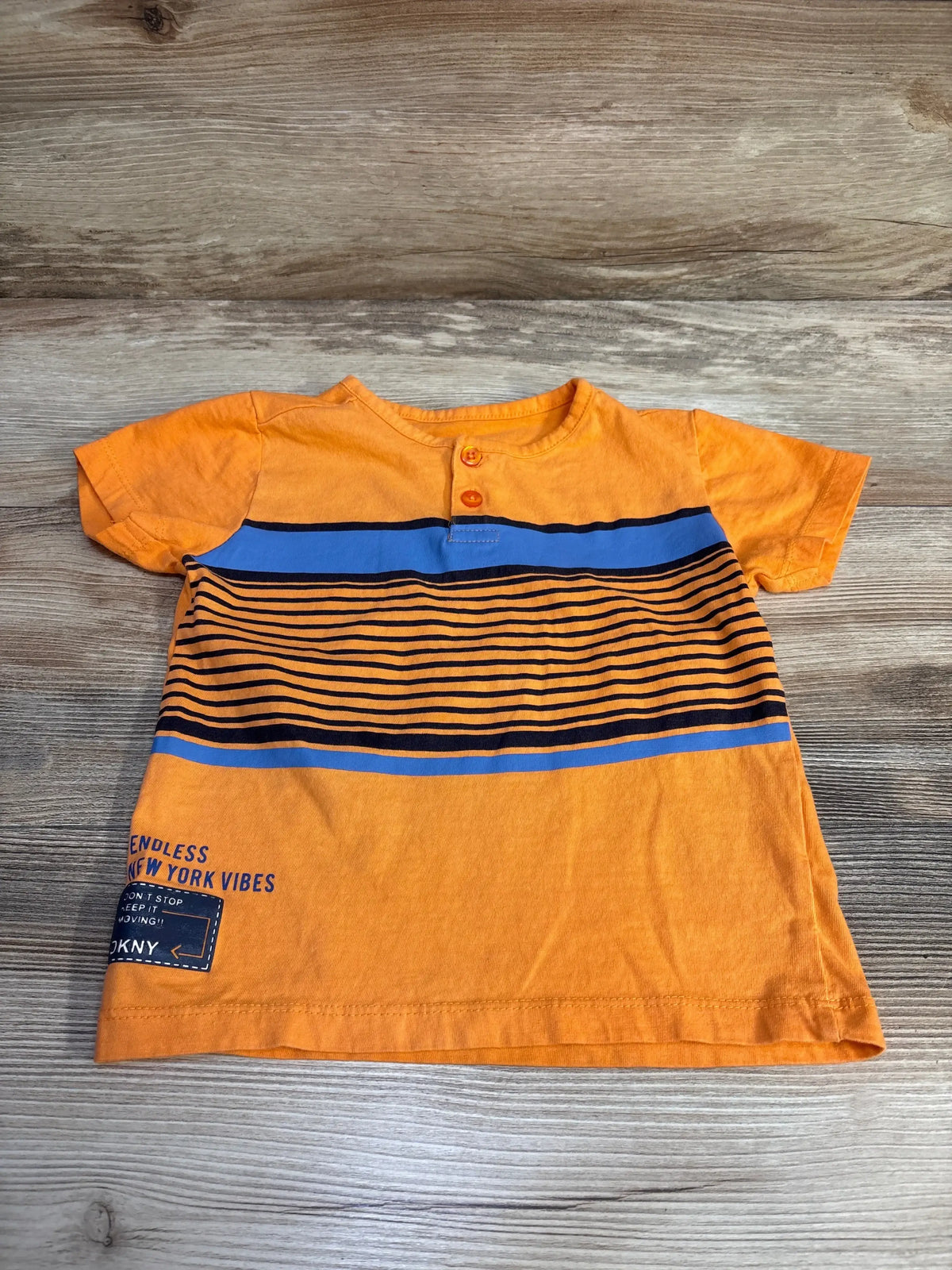 Flat lay of orange DKNY toddler Henley shirt showing blue and black horizontal stripes, two button placket and patch reading Endless New York Vibes on wood background