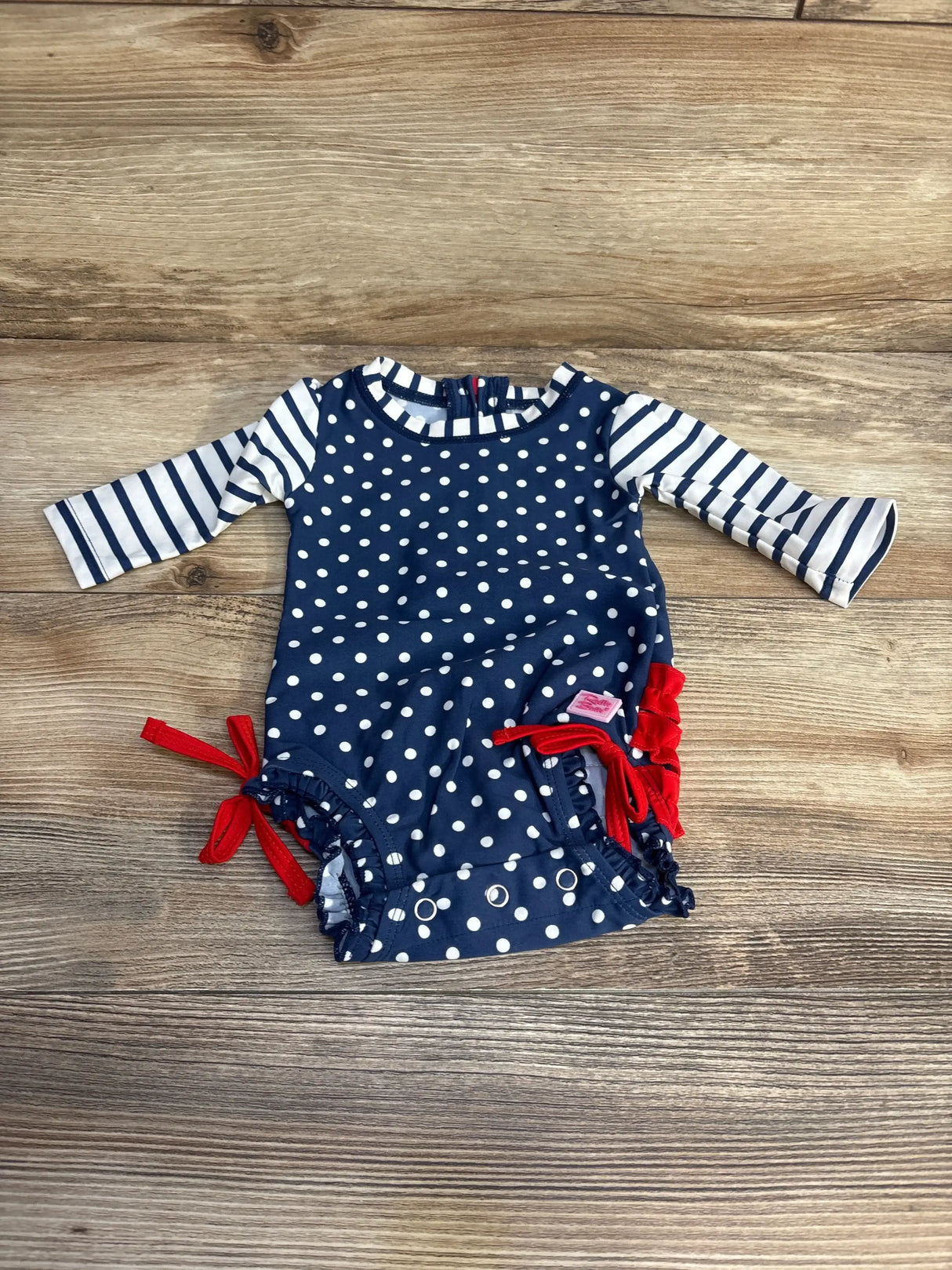 Flat lay of navy baby rashguard one-piece with white polka dots, striped long sleeves, and red side ties on wood background