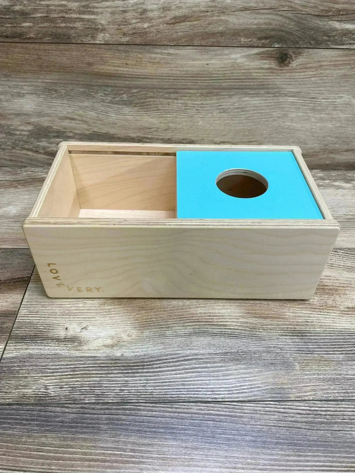 Front view of Lovevery sliding top box showcasing turquoise sliding lid with circular hole on wood plank background