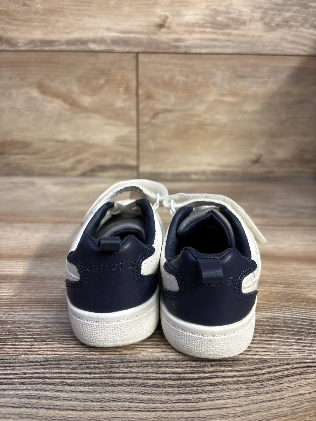 Rear view of Carter's Every Step Port toddler sneakers in white with navy heel panels and Velcro straps on wood-look background