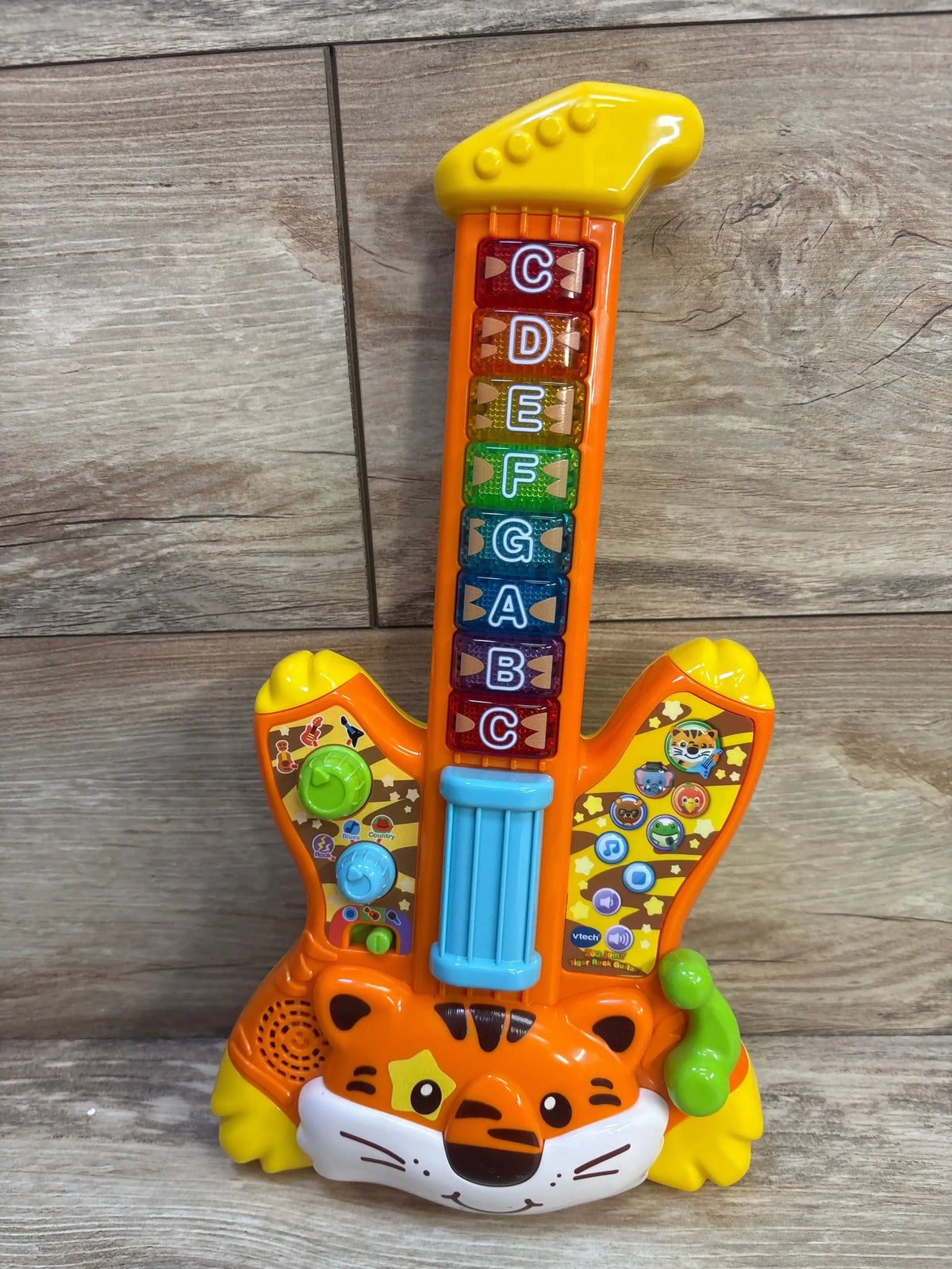 Front view of orange VTech Zoo Jamz Tiger Rock Guitar toy with rainbow letter buttons, blue strum bar, and tiger face base on wood floor
