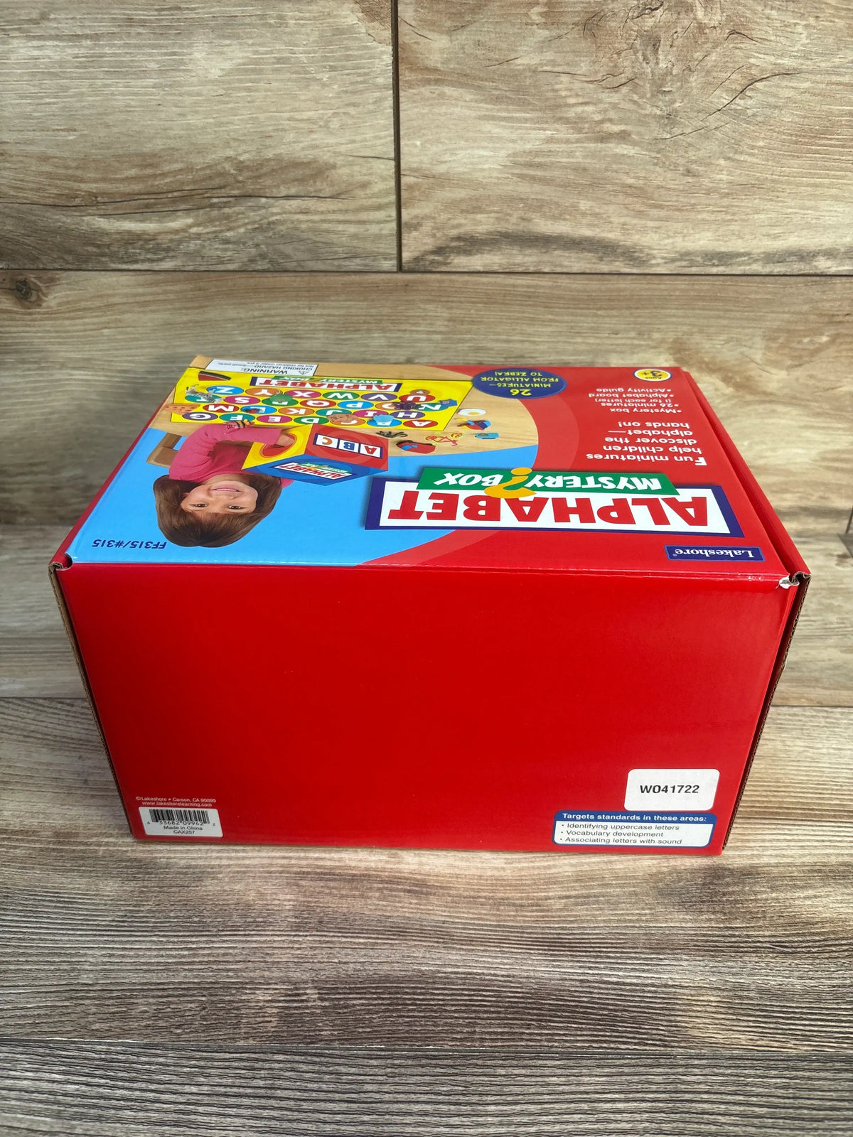 Angled view of Lakeshore Alphabet Mystery Box showcasing alphabet learning graphics on a red box against a wood floor