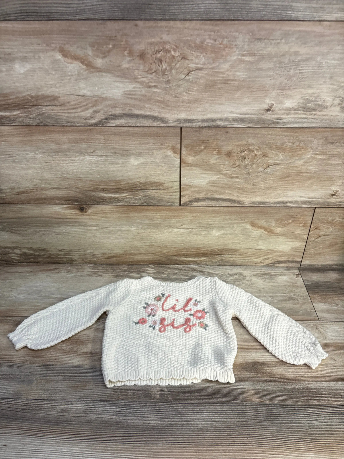 Front view of Max Studio ivory knit sweater in size 18 months showcasing floral Lil Sis embroidery on a wood-grain background
