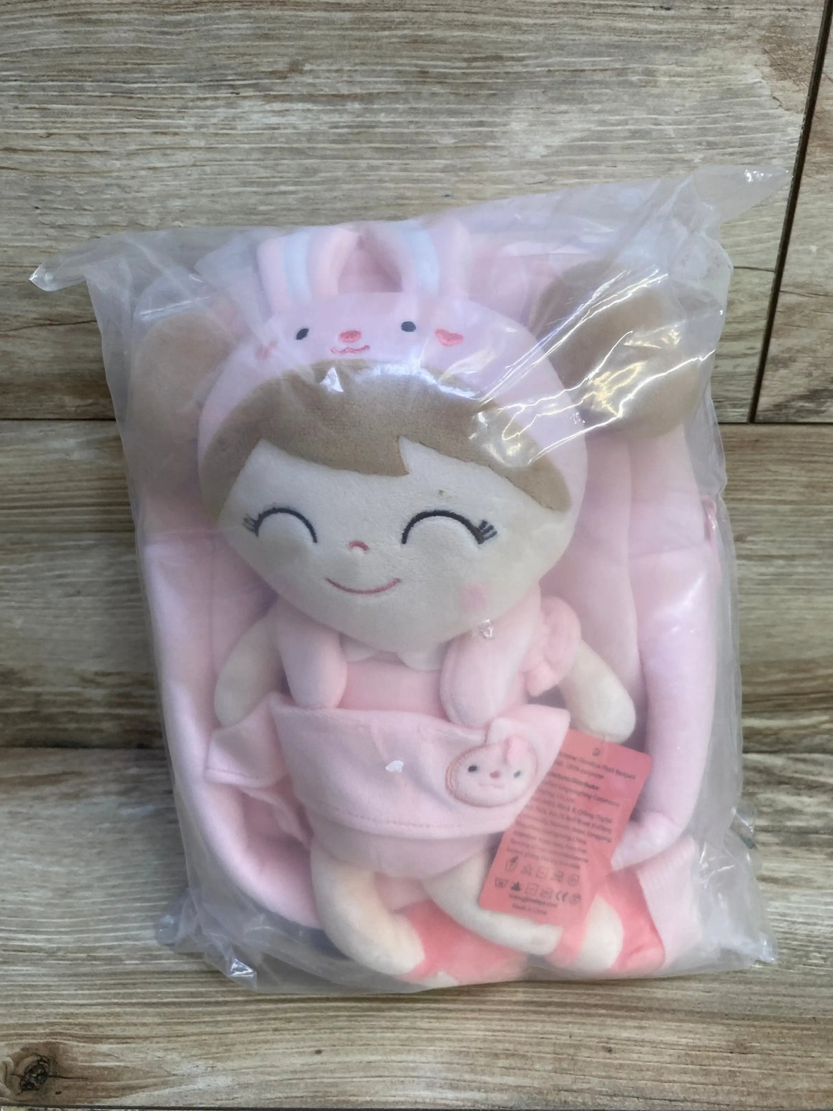 Front view of NEW Gloveleya Pink Rabbit Doll Plush Backpack with bunny-hat doll holding a mini bunny in sealed plastic packaging with hangtag on wood