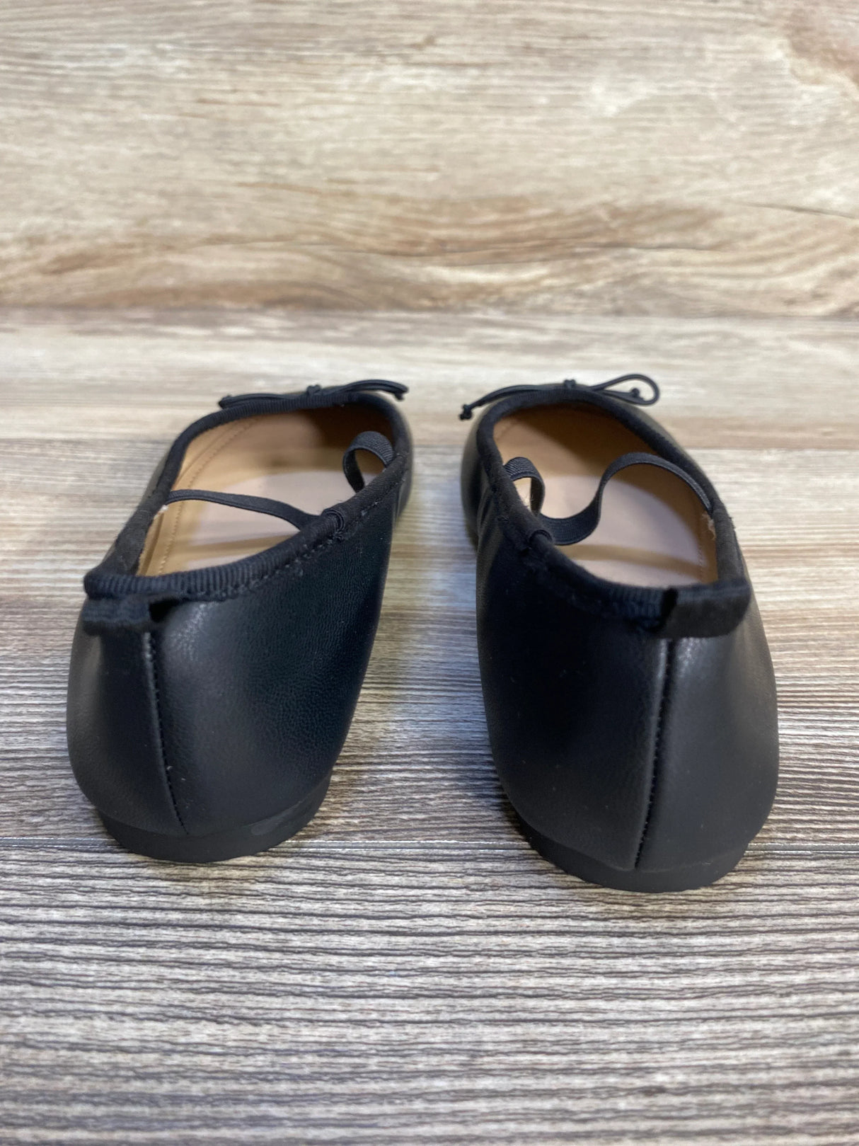 Back view of black toddler ballet flats with elastic straps on wood floor