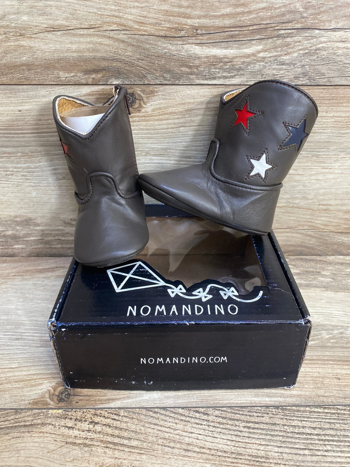 Front view of Nomandino baby boots in chocolate brown leather with red, white, and navy star accents on box, wood background