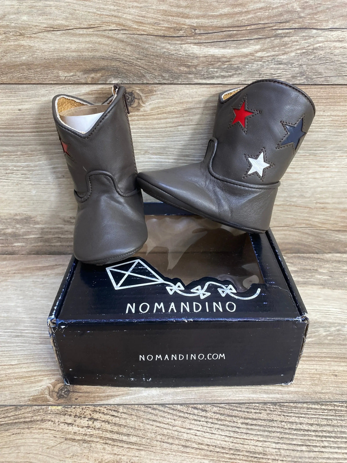 Front view of Nomandino baby boots in chocolate brown leather with red, white, and navy star accents on box, wood background
