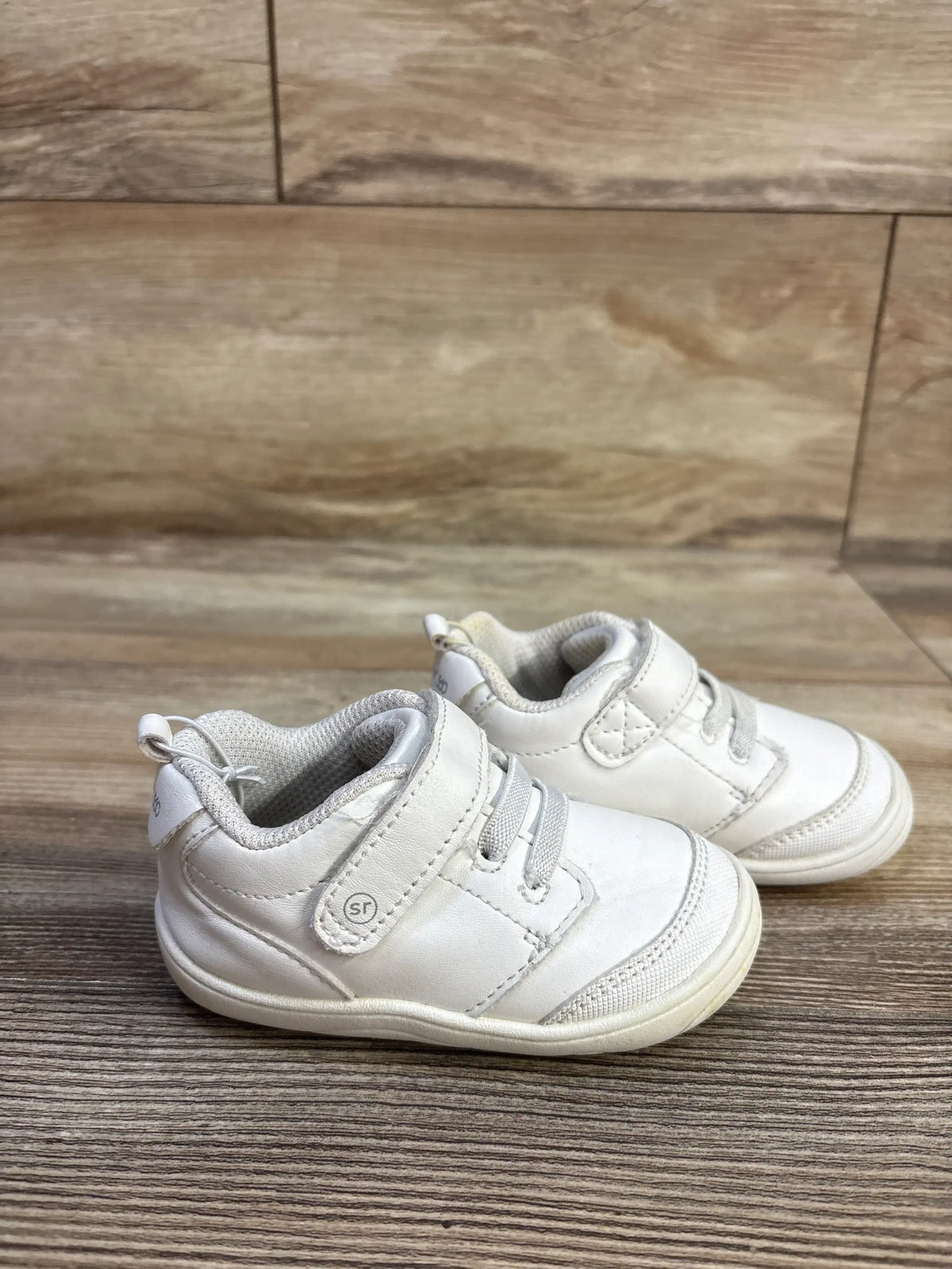 Side view of white Stride Rite Taye 2.0 sneakers in size 5C with lace and hook and loop closures on wood look flooring