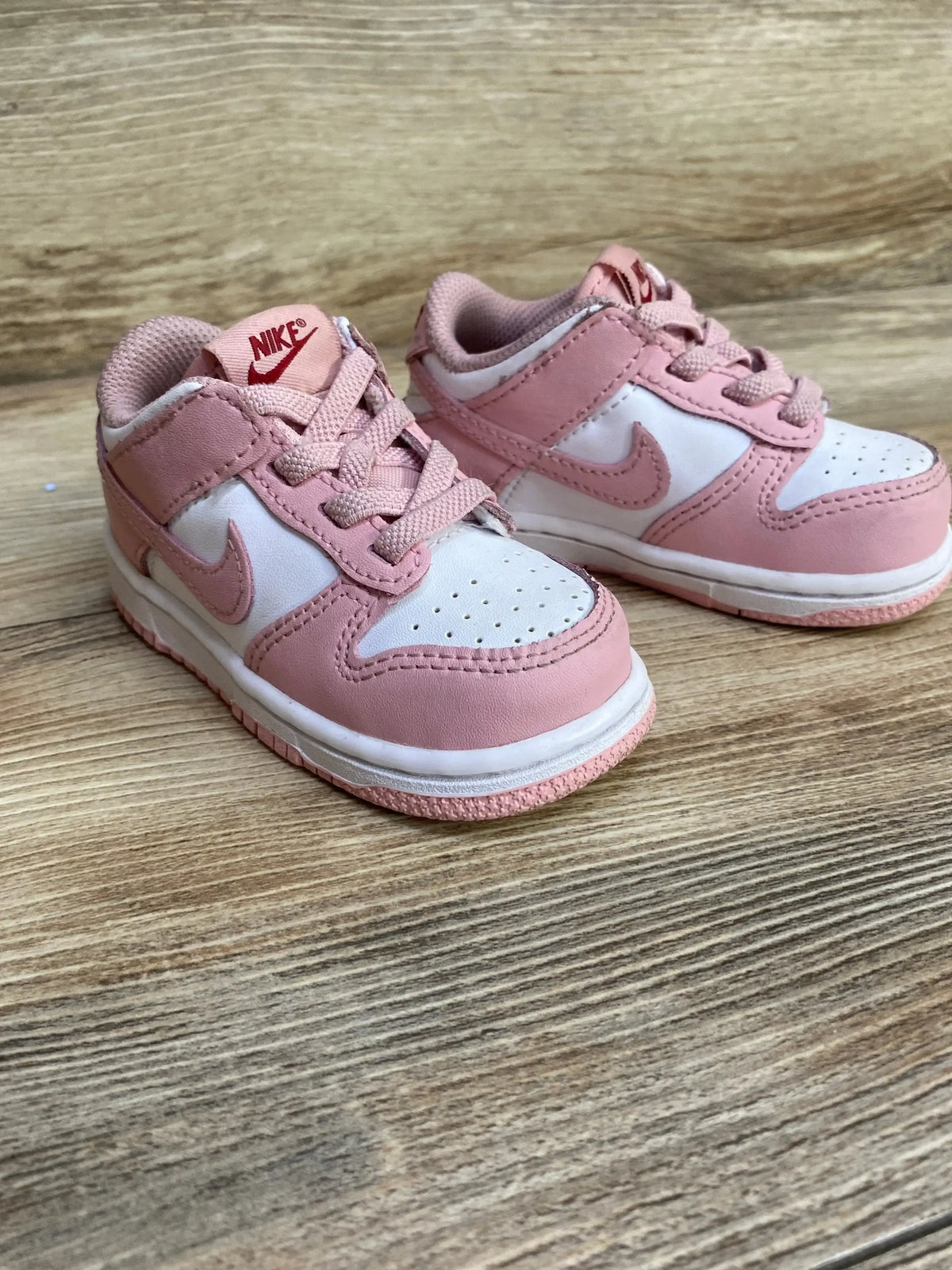 Front-left view of toddler Nike Dunk Low sneakers with pink glaze overlays, white panels and laces on wood surface, size 6C