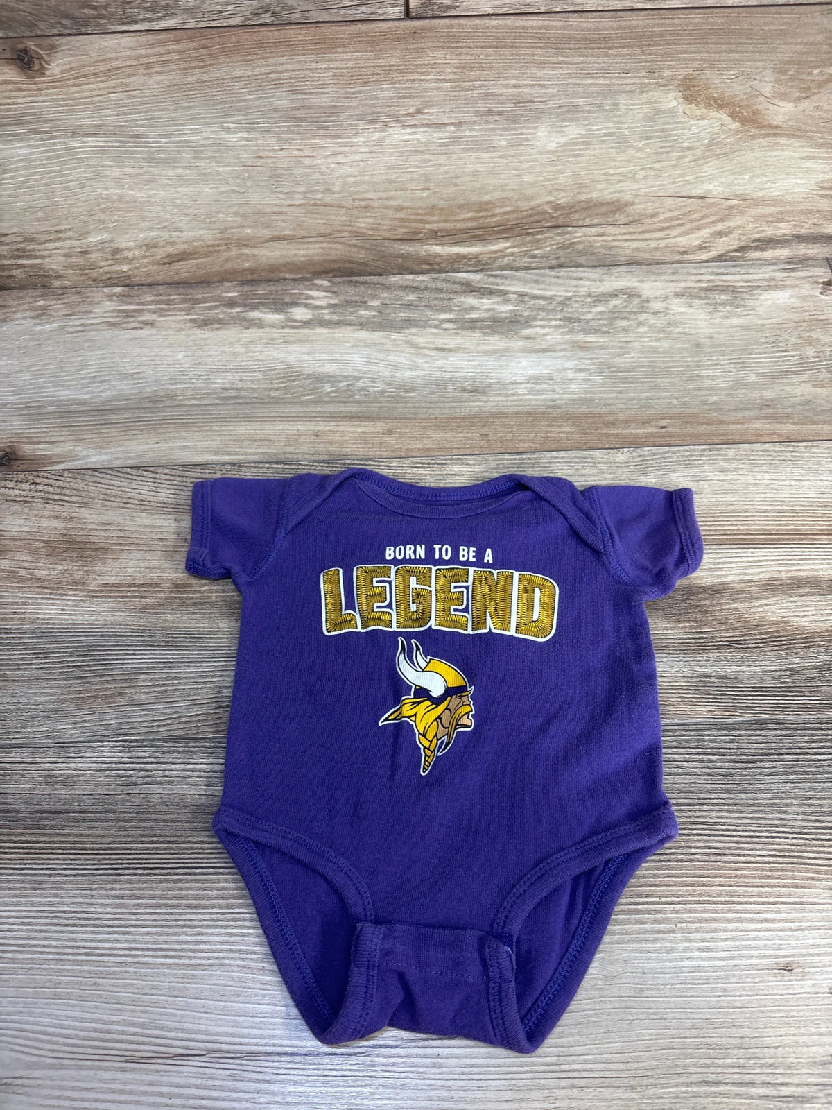 Front view of purple Minnesota Vikings bodysuit showcasing Born to Be a Legend graphic on wood-grain background