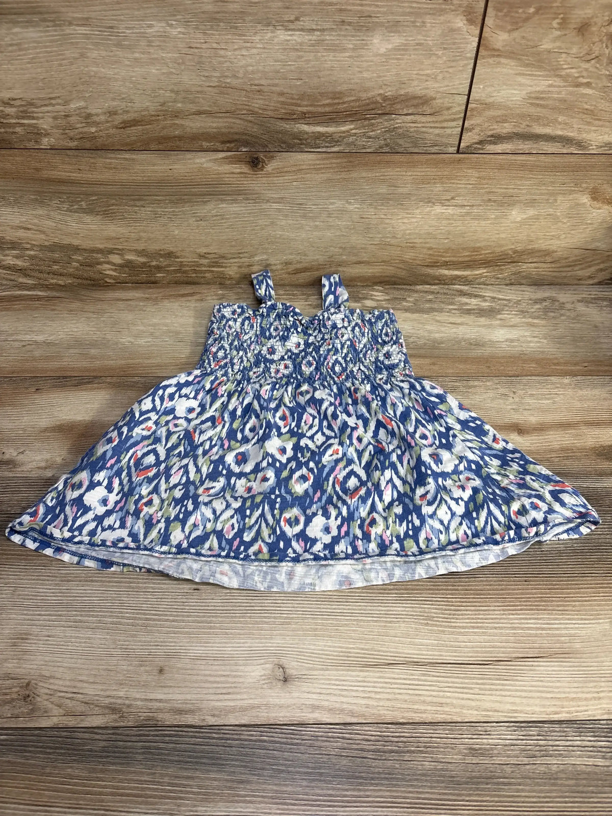Flat lay of blue Oshkosh smocked toddler dress with smocked bodice, thin straps, and multicolor print on wood