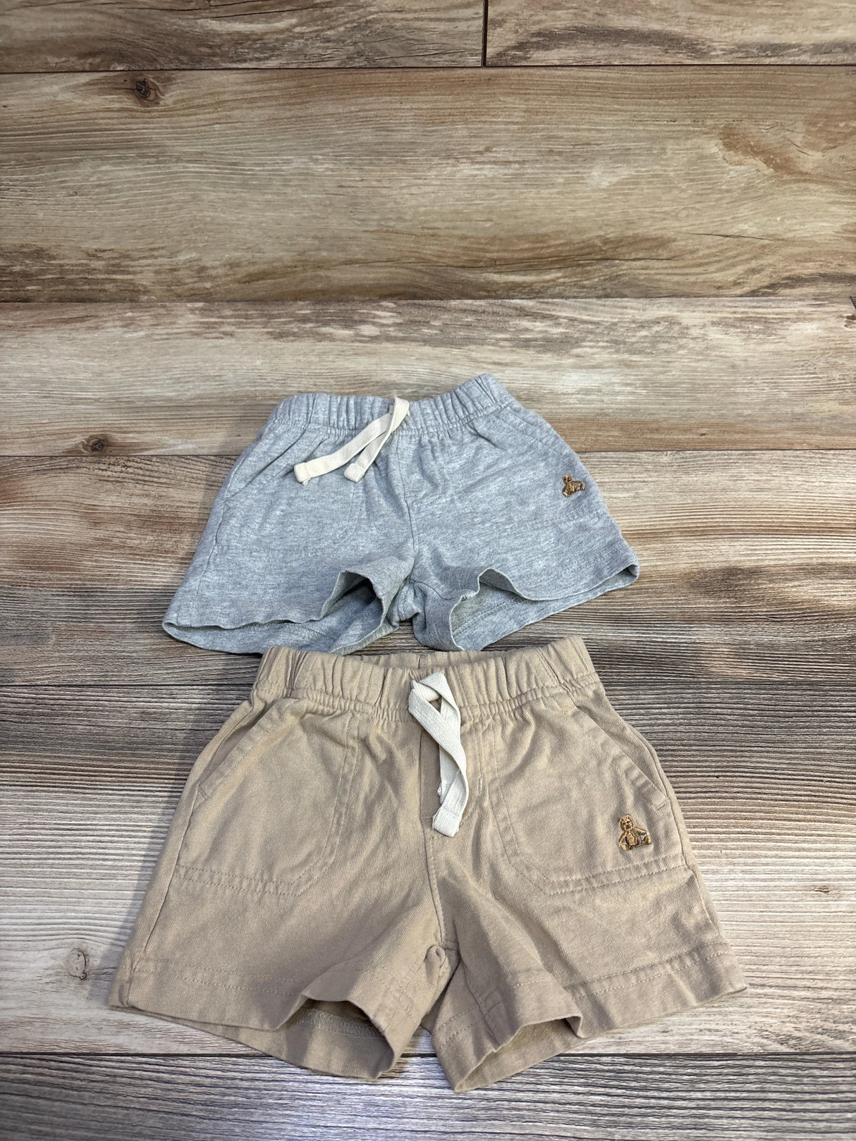 Top-down view of two Baby Gap pull-on baby shorts showcasing elastic waistbands, drawstrings, pockets, and embroidered teddy logos on a wood floor background
