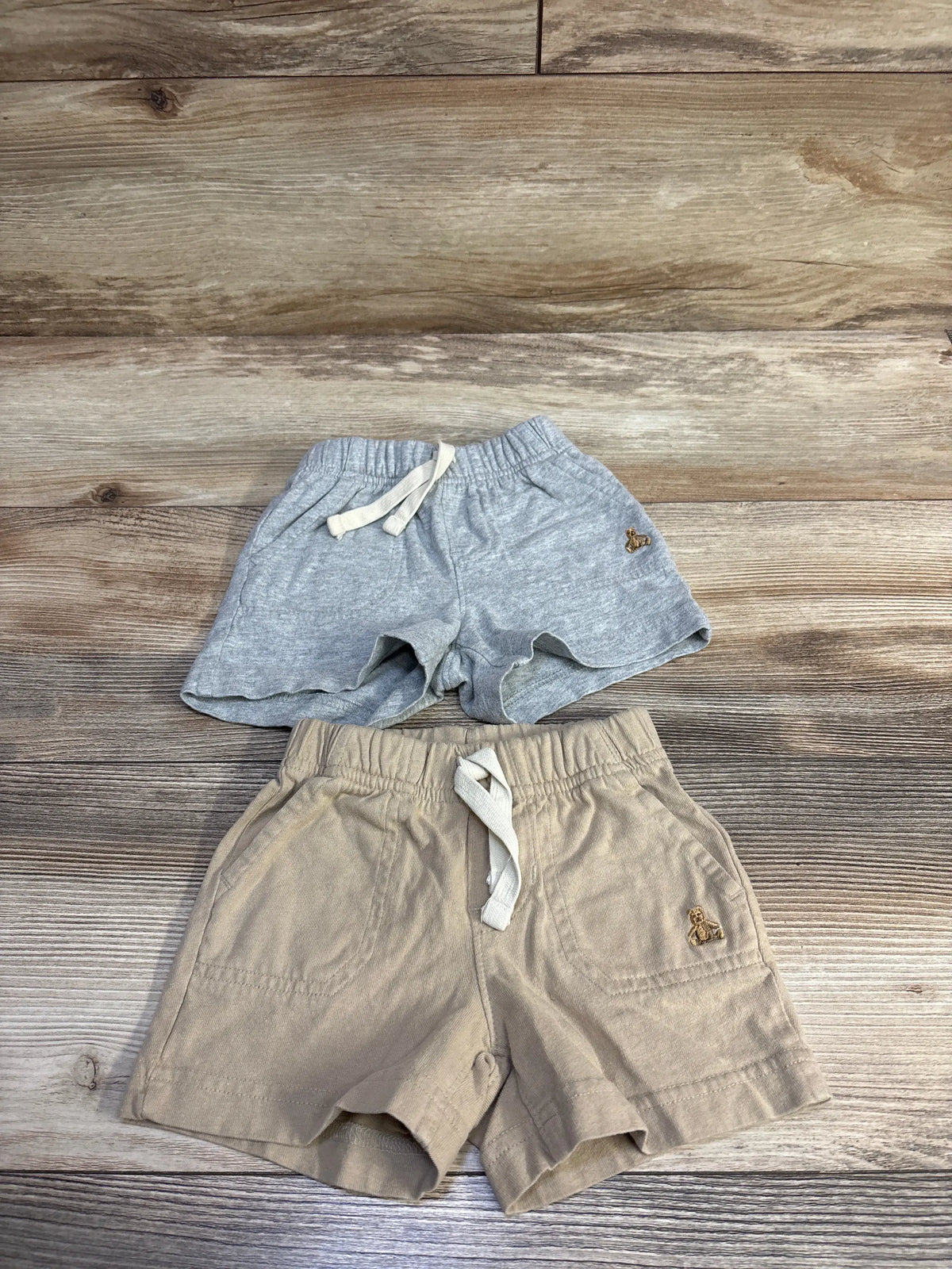 Top-down view of two Baby Gap pull-on baby shorts showcasing elastic waistbands, drawstrings, pockets, and embroidered teddy logos on a wood floor background