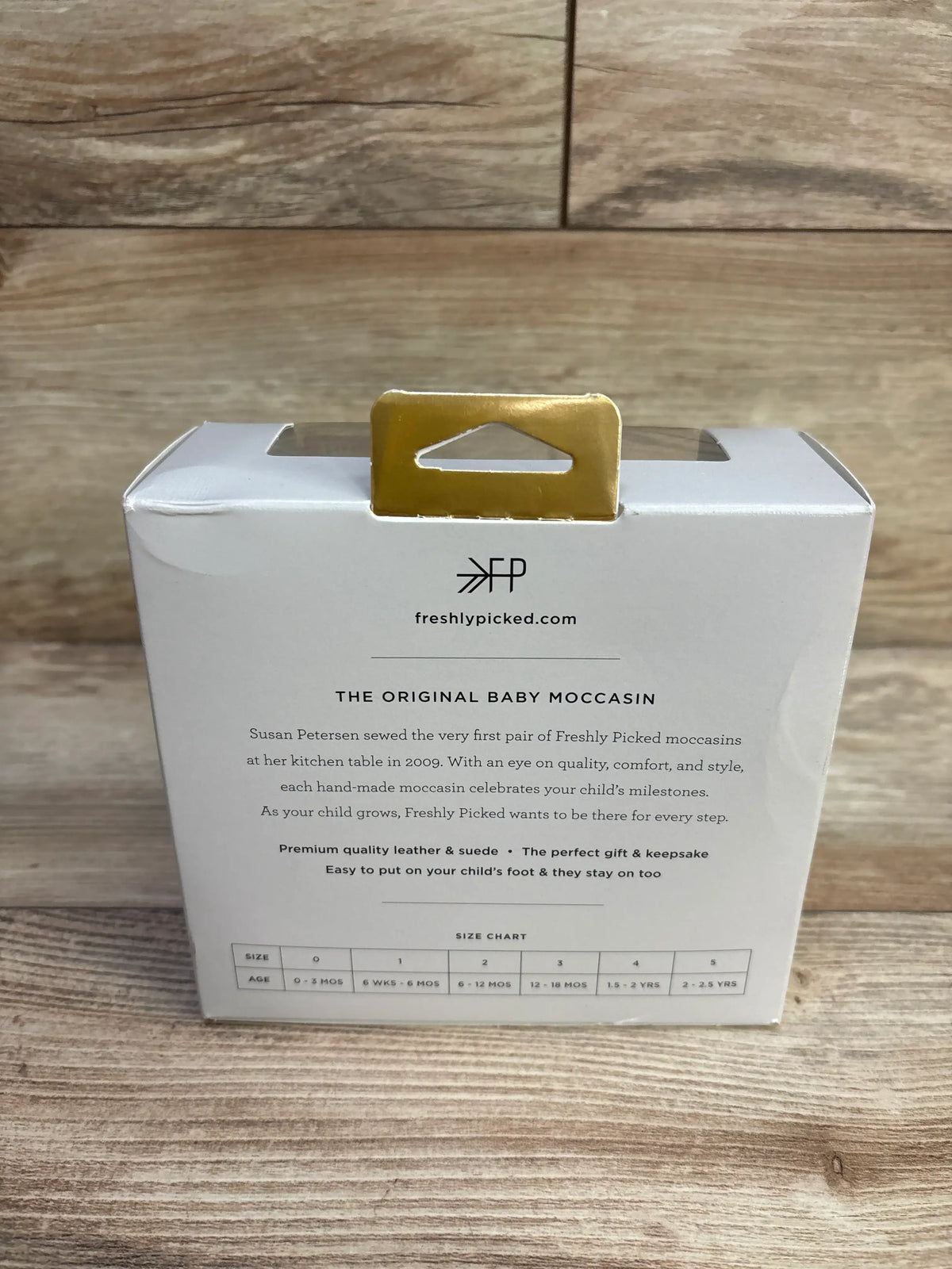 Rear view of Freshly Picked Tropical baby moccasin box showing brand logo, product description and size chart on wood background