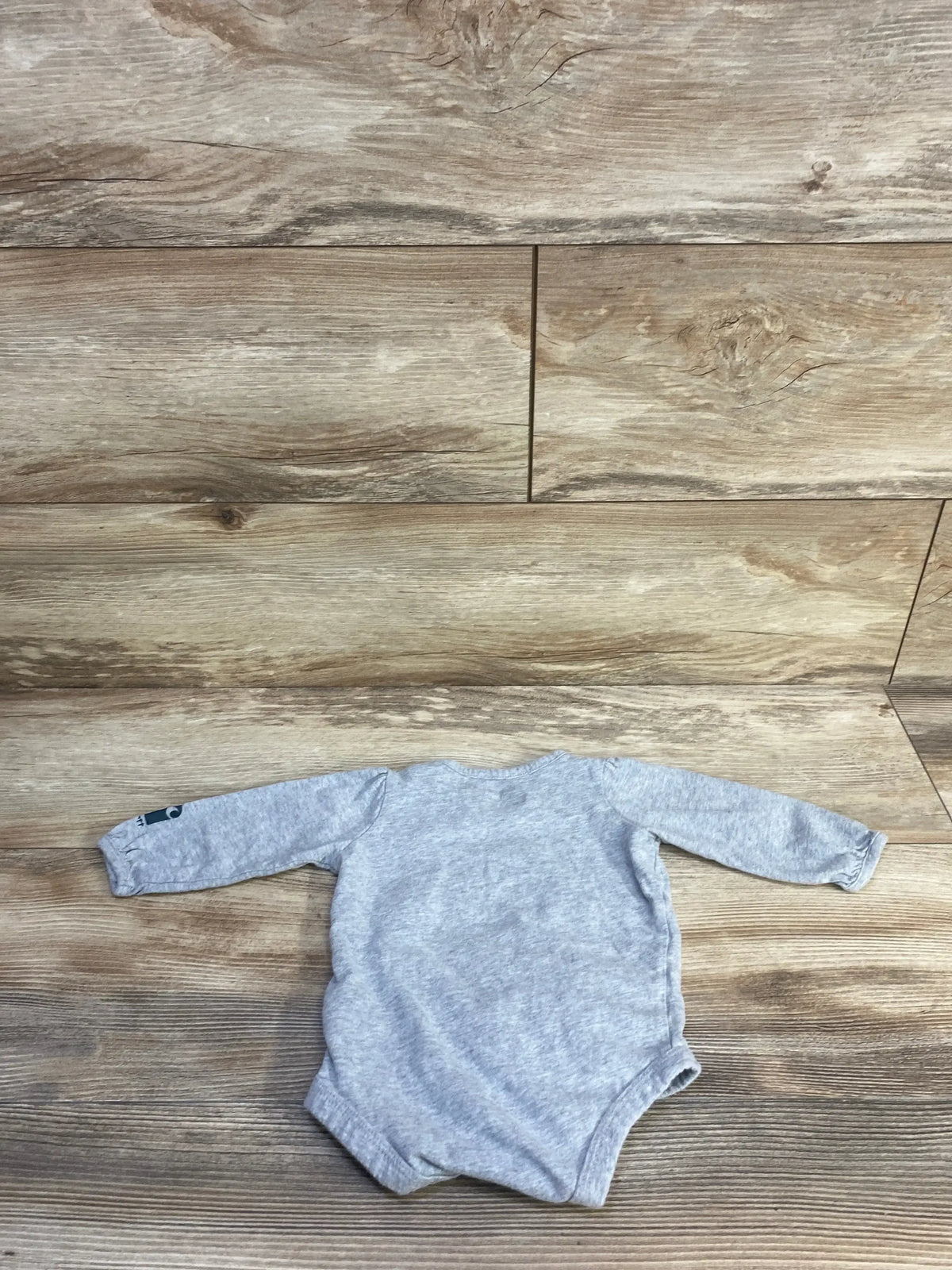 Back view of grey Carhartt long-sleeve baby bodysuit (6 months) showing small sleeve logo on wood floor background