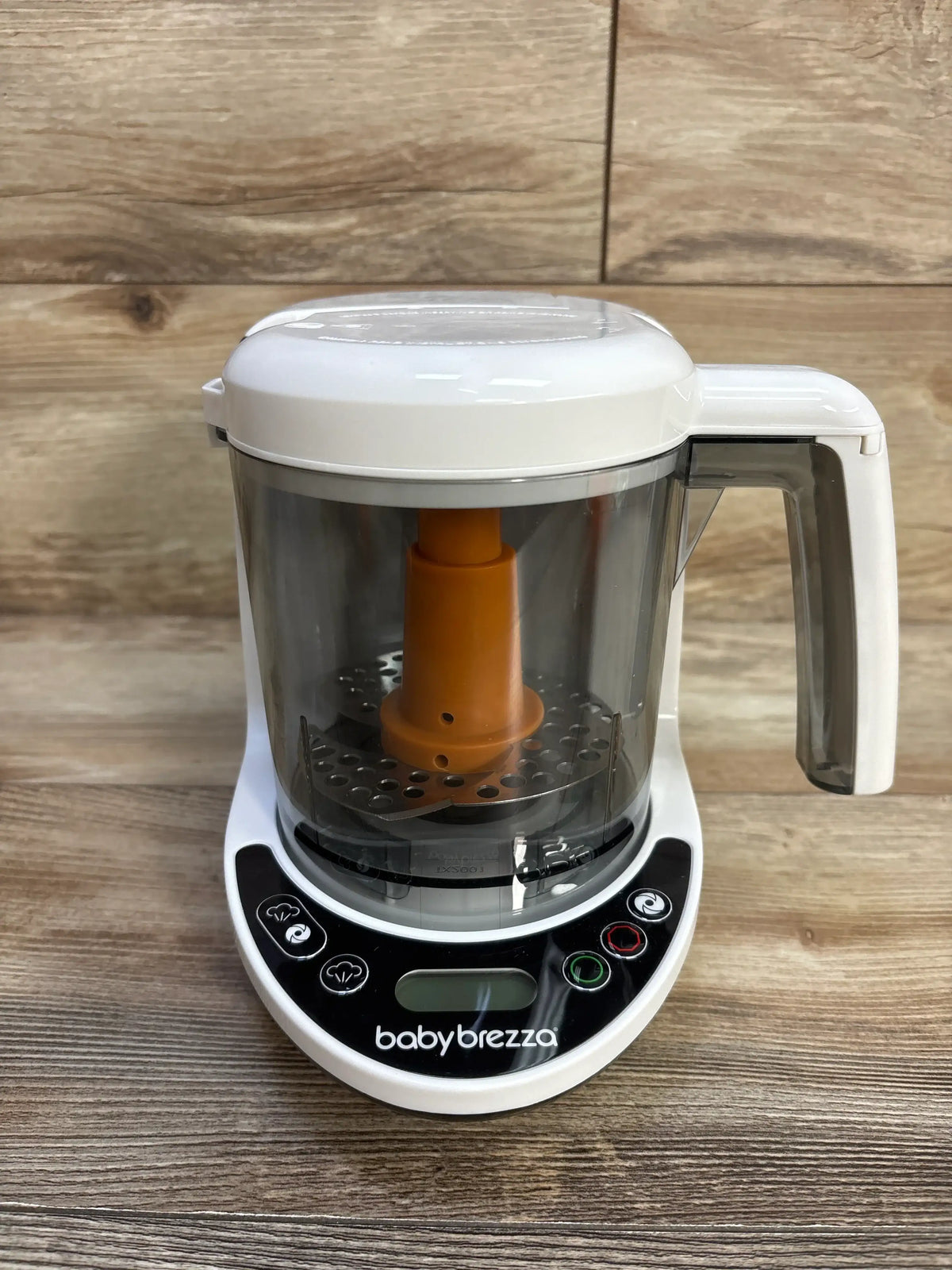 Front view of Baby Brezza One Step Baby Food Maker Deluxe with clear bowl, orange blade assembly, and control panel on wood