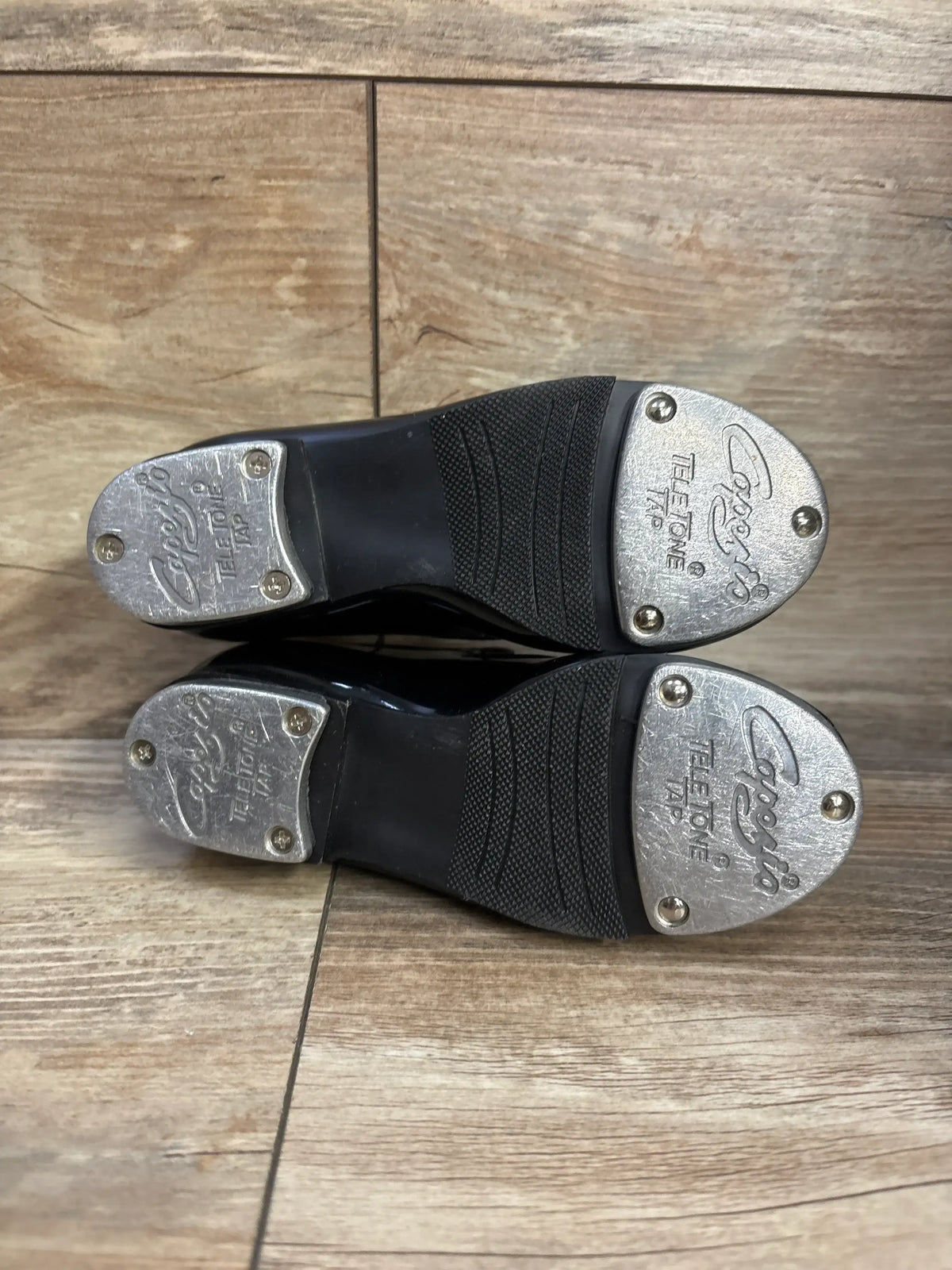 Bottom view of Capezio TeleTone tap shoes in black, size 8.5C showcasing metal TeleTone taps and textured rubber sole on wood floor