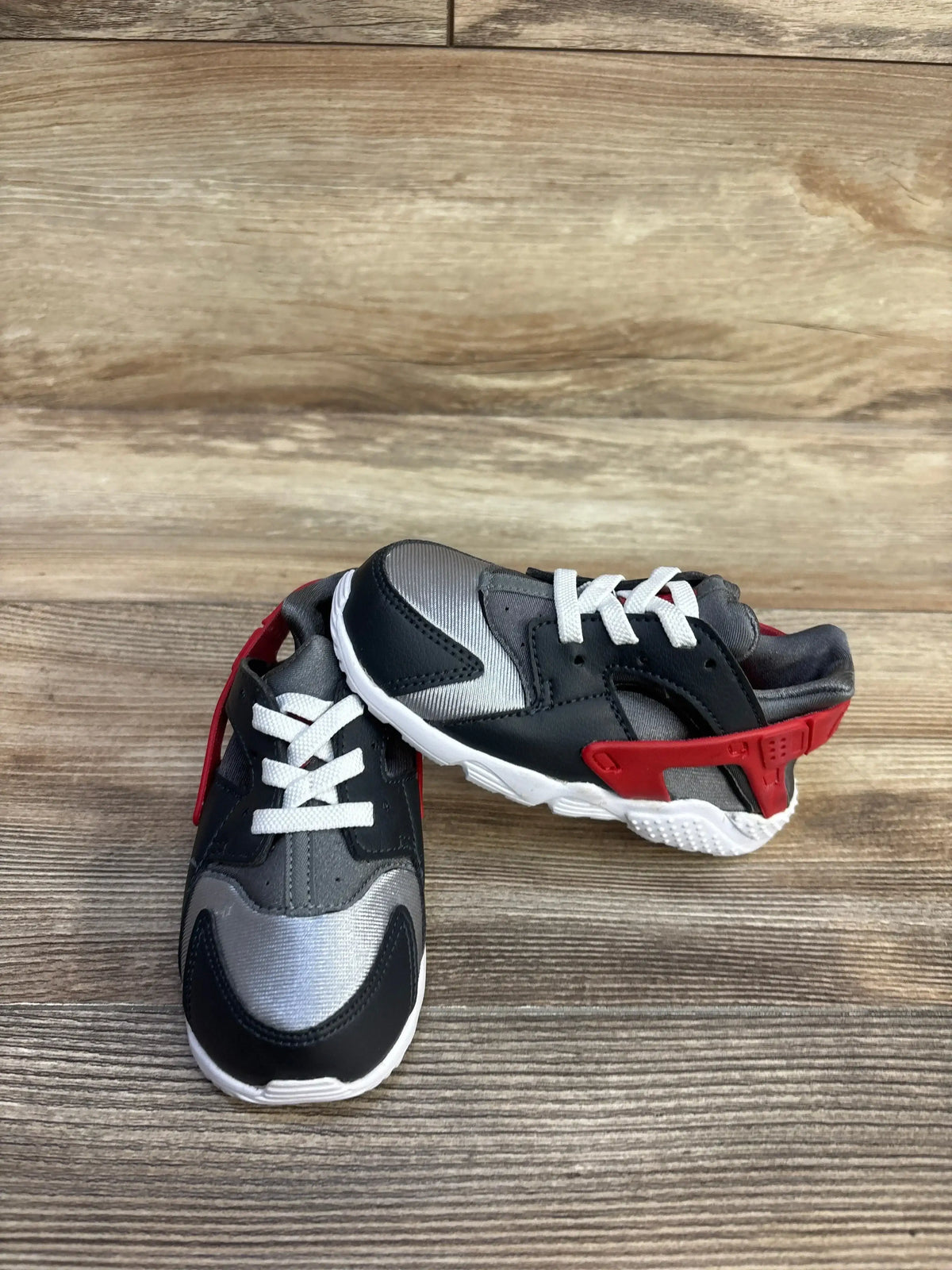 Three-quarter view of Nike Huarache Run TD sneakers in dark smoke gray with silver panels and university red heel cage on wood plank floor