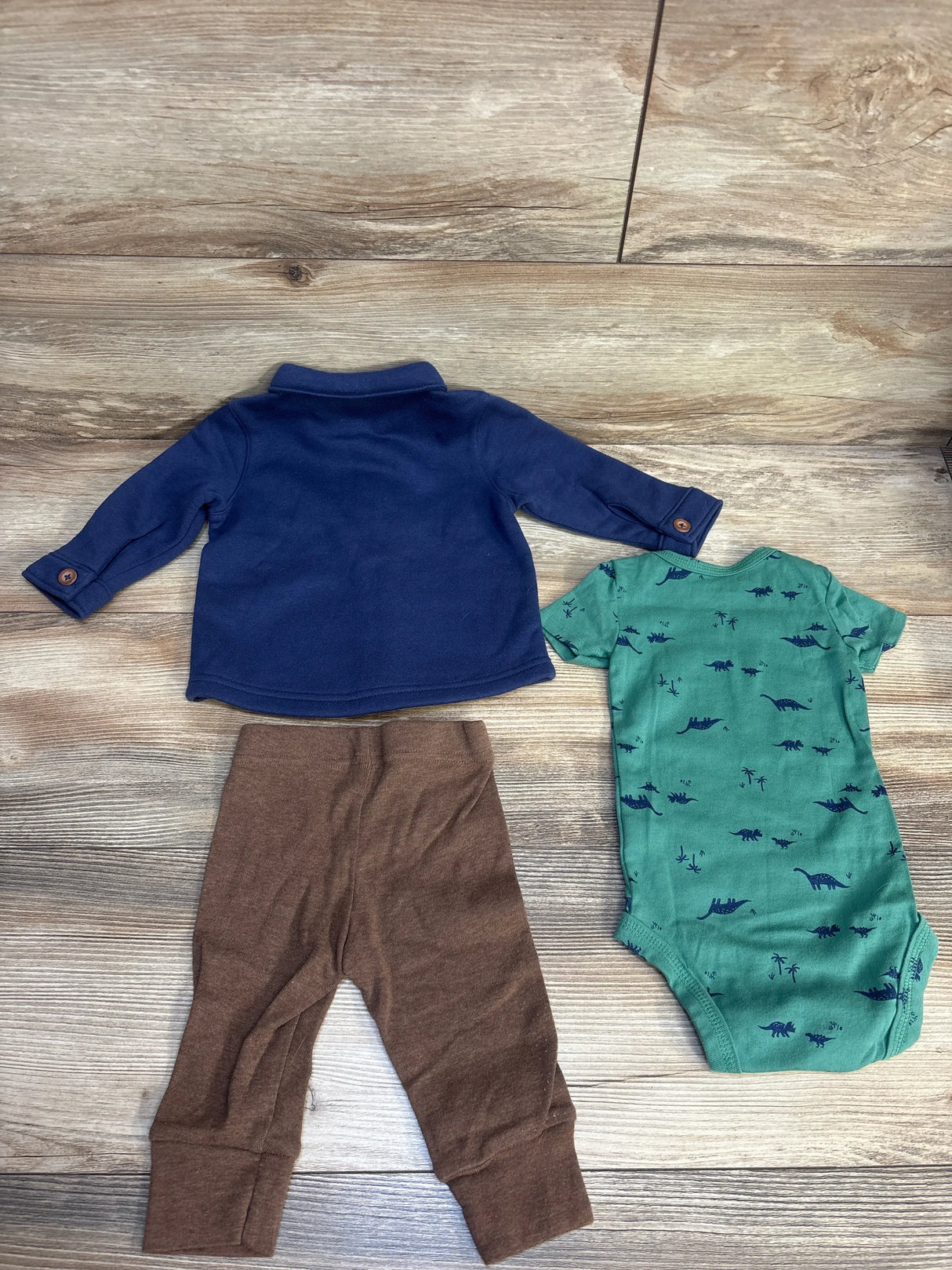 Top-down view of Just One You 3pc Set showcasing navy jacket, brown joggers, and teal dinosaur-print bodysuit on wood flooring