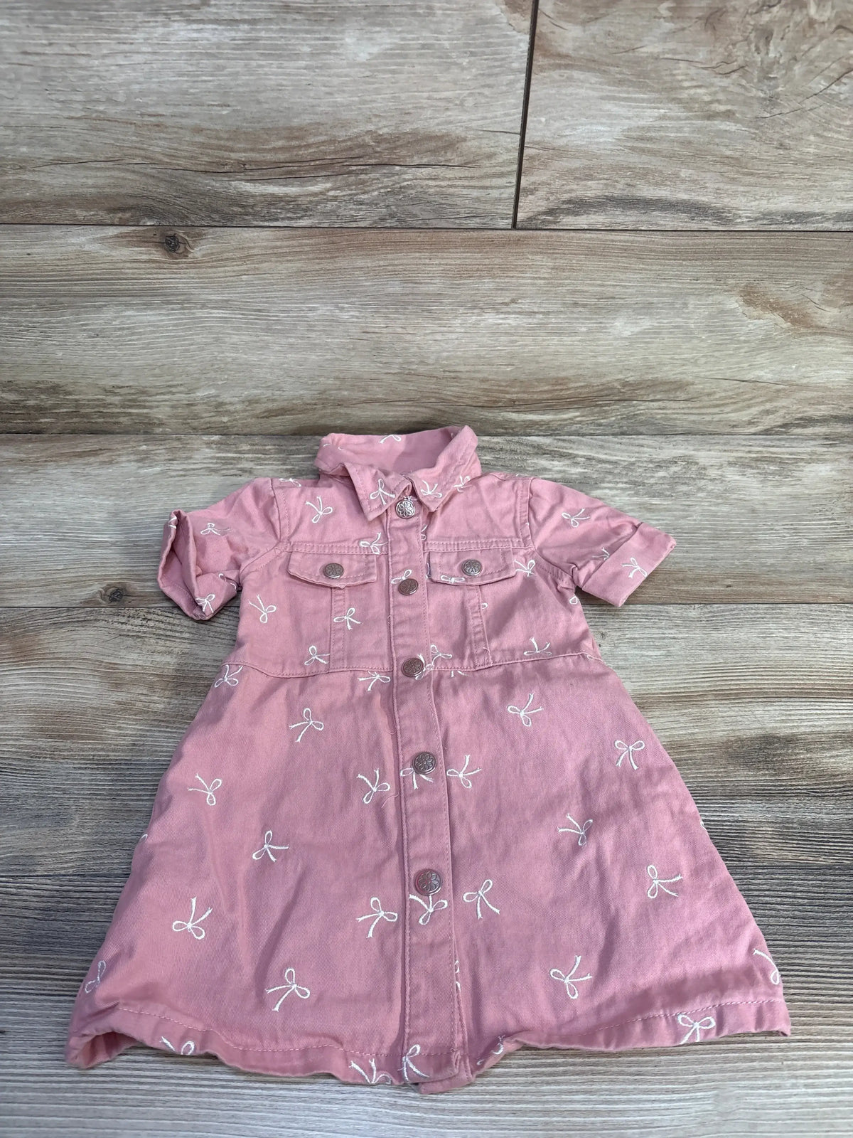 Flat lay of girls’ Nanette button-up dress in pink with front buttons and white embroidered bows on wood background