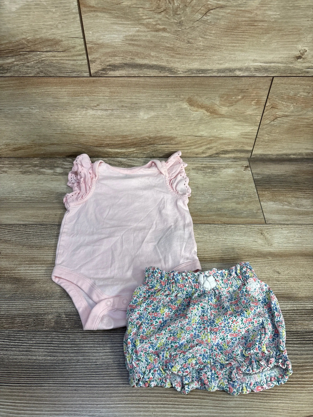Top view of Baby Gap bodysuit and shorts set showcasing a pink bodysuit, floral ruffled shorts, and eyelet trim on wood flooring