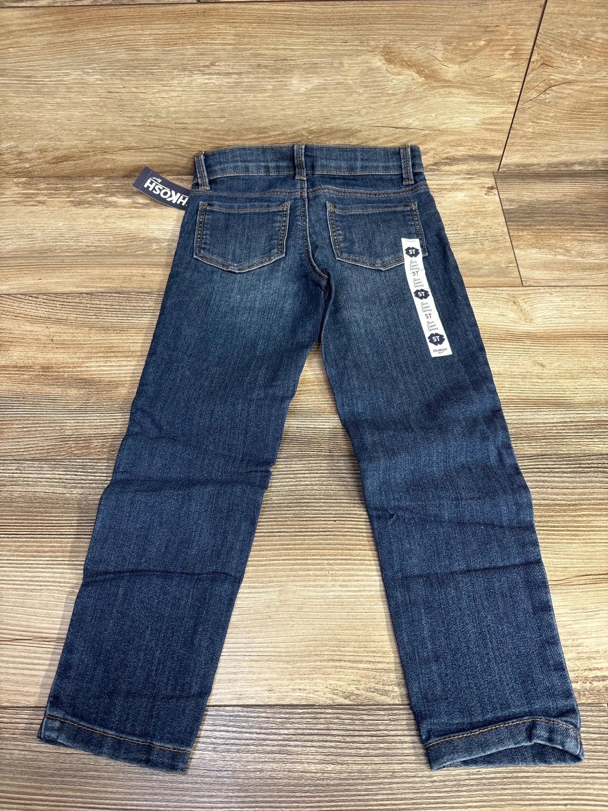 Flat lay of OshKosh Super Skinny toddler jeans size 5T showing dark blue wash, back pockets and retailer tag on wood floor